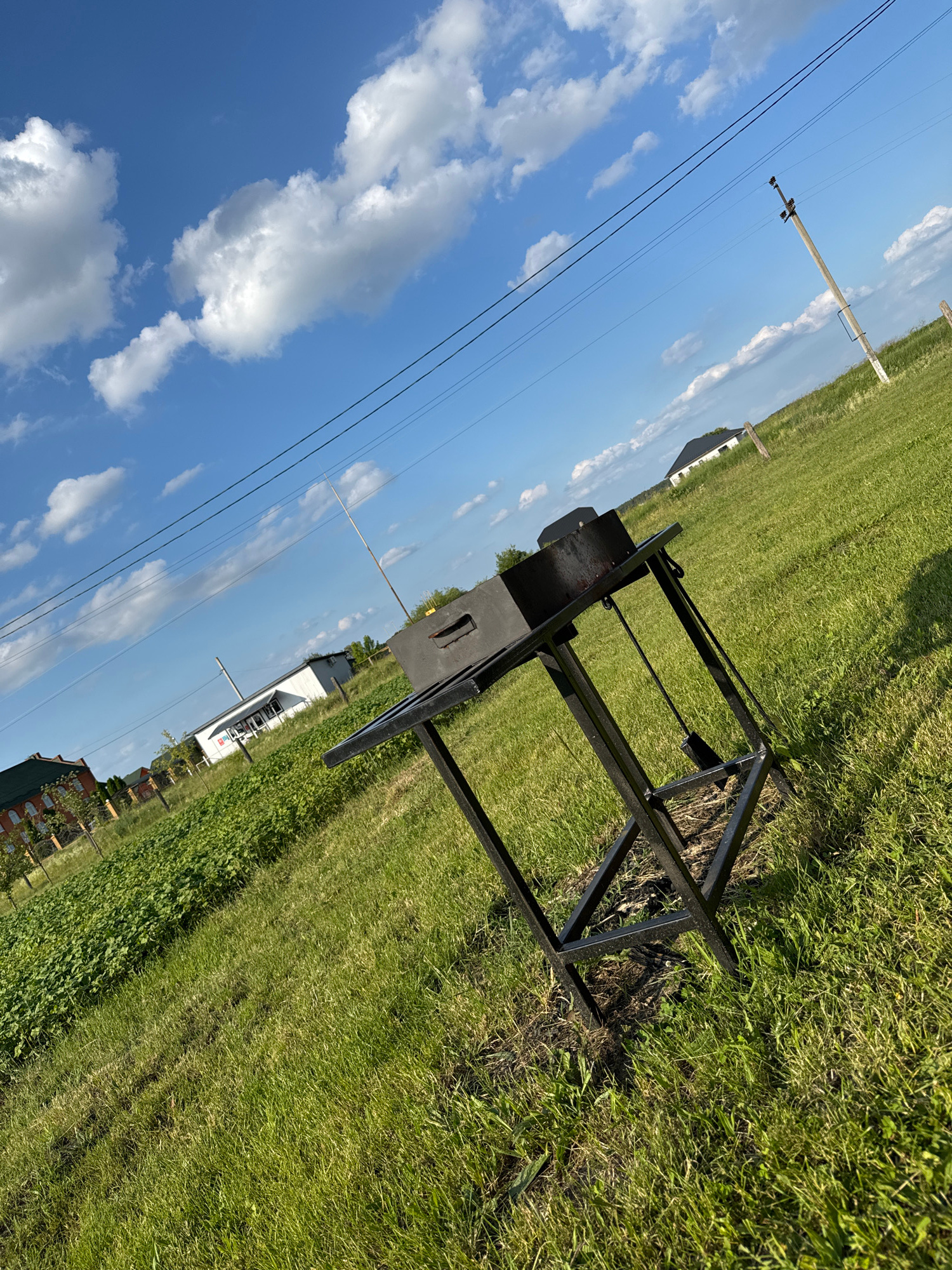 This image captures an outdoor scene under a bright blue sky with scattered white clouds. The perspective is tilted, giving a dynamic feel to the composition. In the foreground, a black metal structure with a rectangular base and an X-shaped support system is set on a grassy lawn. This object appears to be some sort of equipment or stand.
Further in the background, a utility pole with wires stretches diagonally across the frame, suggesting a rural or semi-rural setting. Beyond that, a white building with a dark roof is visible, possibly a house or another structure. Adjacent to the building, a patch of lush green vegetation, possibly a garden, can be seen. In the far distance, the roofs of more buildings are peeking out, hinting at a nearby settlement.
The scene appears to be set during the daytime, likely late morning or afternoon, given the clear sky and the sunlight casting shadows. The weather is fair and sunny. The overall impression is one of a quiet, everyday moment in a Ukrainian landscape. No people are visible in the image. There is no discernible text in the image.