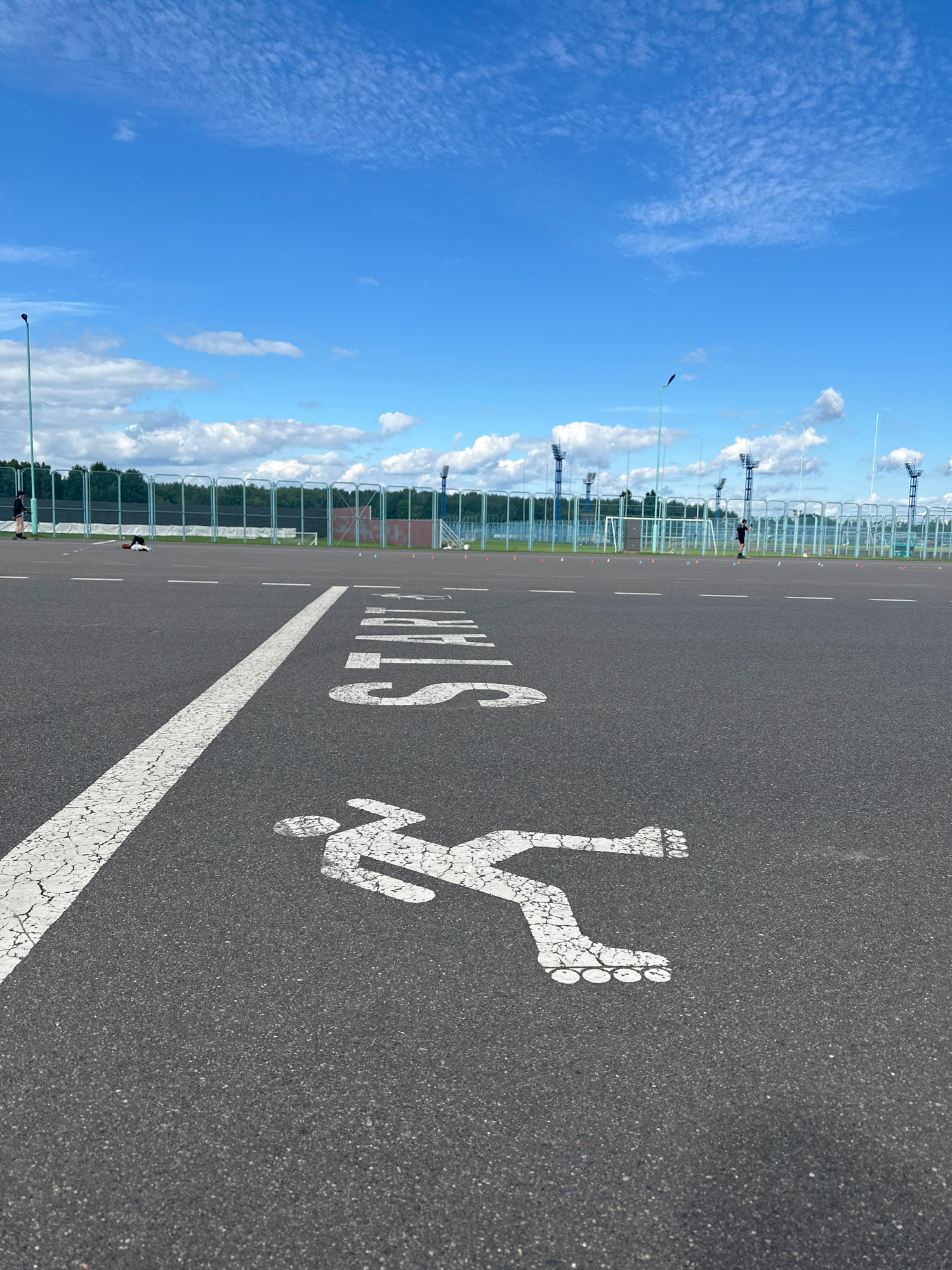 The image shows a rollerblading track on an asphalt surface. A white line marking the lane is cracked and faded. A white stencil of a rollerblader is painted on the track, facing to the left. The word "START" is also painted in white letters on the track. In the background, a fence separates the track from a grassy area with trees. Several lampposts are visible, suggesting an outdoor sports facility. The sky is bright blue with scattered white clouds. The orientation of the image is vertical, making it appear as if the viewer is looking down at an angle. There are no people clearly visible in the image. The time of day appears to be daytime, likely morning or afternoon, given the bright sunlight. The weather seems to be clear and pleasant.