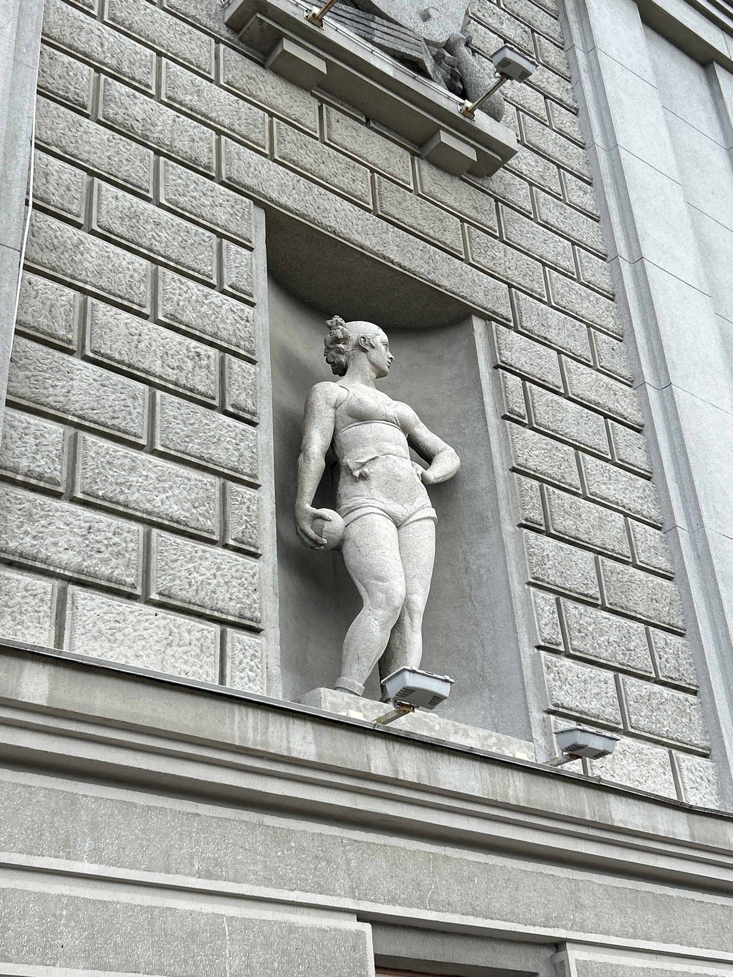 The image shows a stone sculpture of a nude athletic figure holding a ball, set in a recessed niche on the facade of a building. The building's exterior is clad in stone blocks that are textured and arranged in a regular pattern. The sculpture depicts a muscular figure with short, curly hair, standing with one hand on its hip and the other holding a ball at its side. The niche is rectangular and deep, with the sculpture positioned towards the front. Two rectangular lights with grey casings are mounted on brackets below the niche, projecting outwards. The surrounding building structure is made of grey stone and features horizontal banding and recessed sections. The overall impression is of classical architecture with a modernist sculptural element. The building is likely a public or institutional building, given the scale and style of the sculpture. The location is Novokuybyshevsk, Russia.