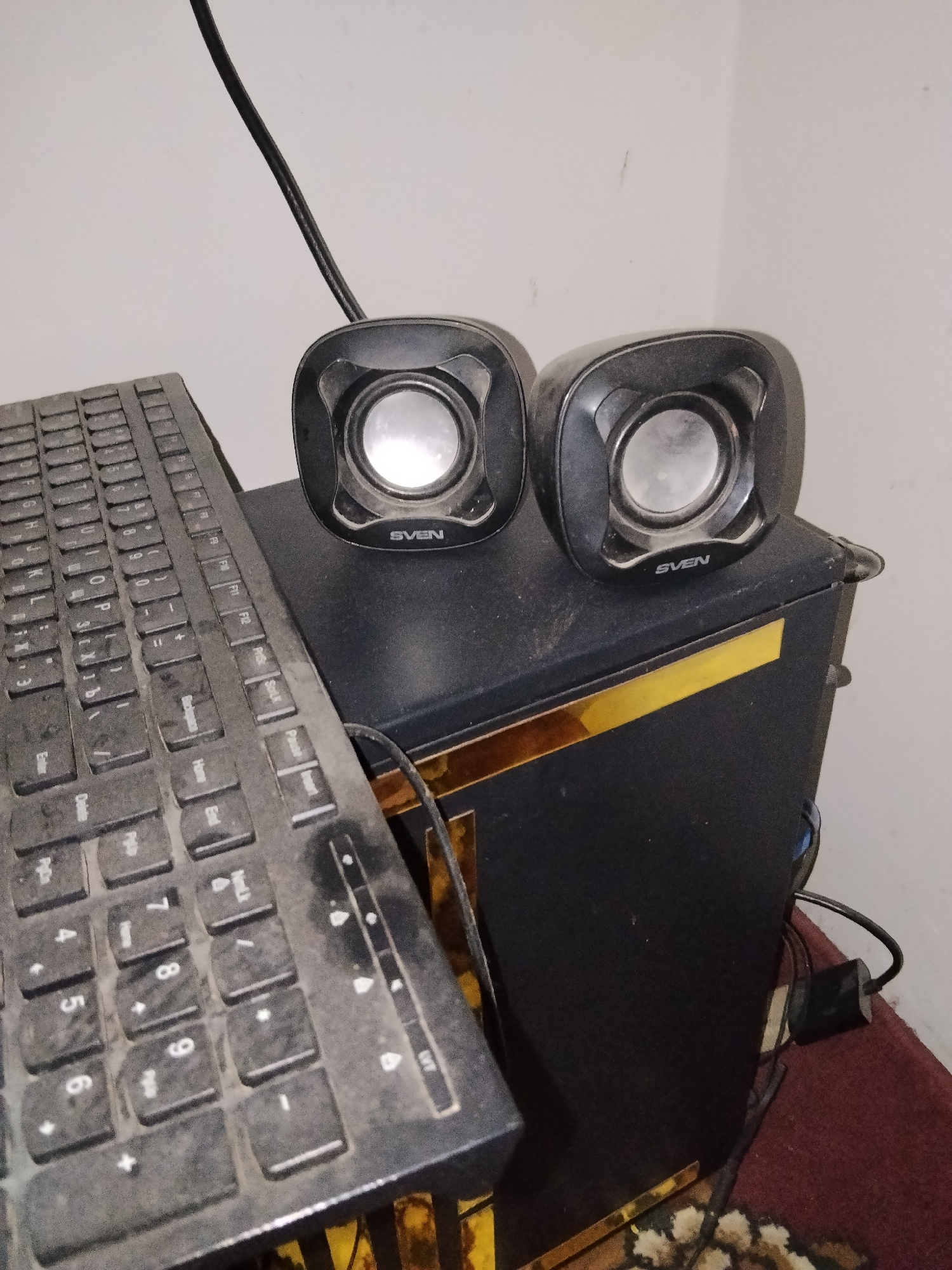 A black computer keyboard is positioned in the foreground, angled from the lower left to the upper left of the image. Two small, black speakers, each with a silver circular speaker cone, are placed on top of a black subwoofer. The subwoofer has a gold trim line across its front. A black cable runs from behind the speakers and subwoofer, disappearing out of frame to the left. The background consists of a plain, light-colored wall. The scene is lit from an unseen source, casting subtle shadows. The image is cropped and slightly angled, suggesting it was taken from a standing or slightly elevated position. The setting appears to be a home or office environment within Yangibazar, Uzbekistan, where the computer equipment is set up.