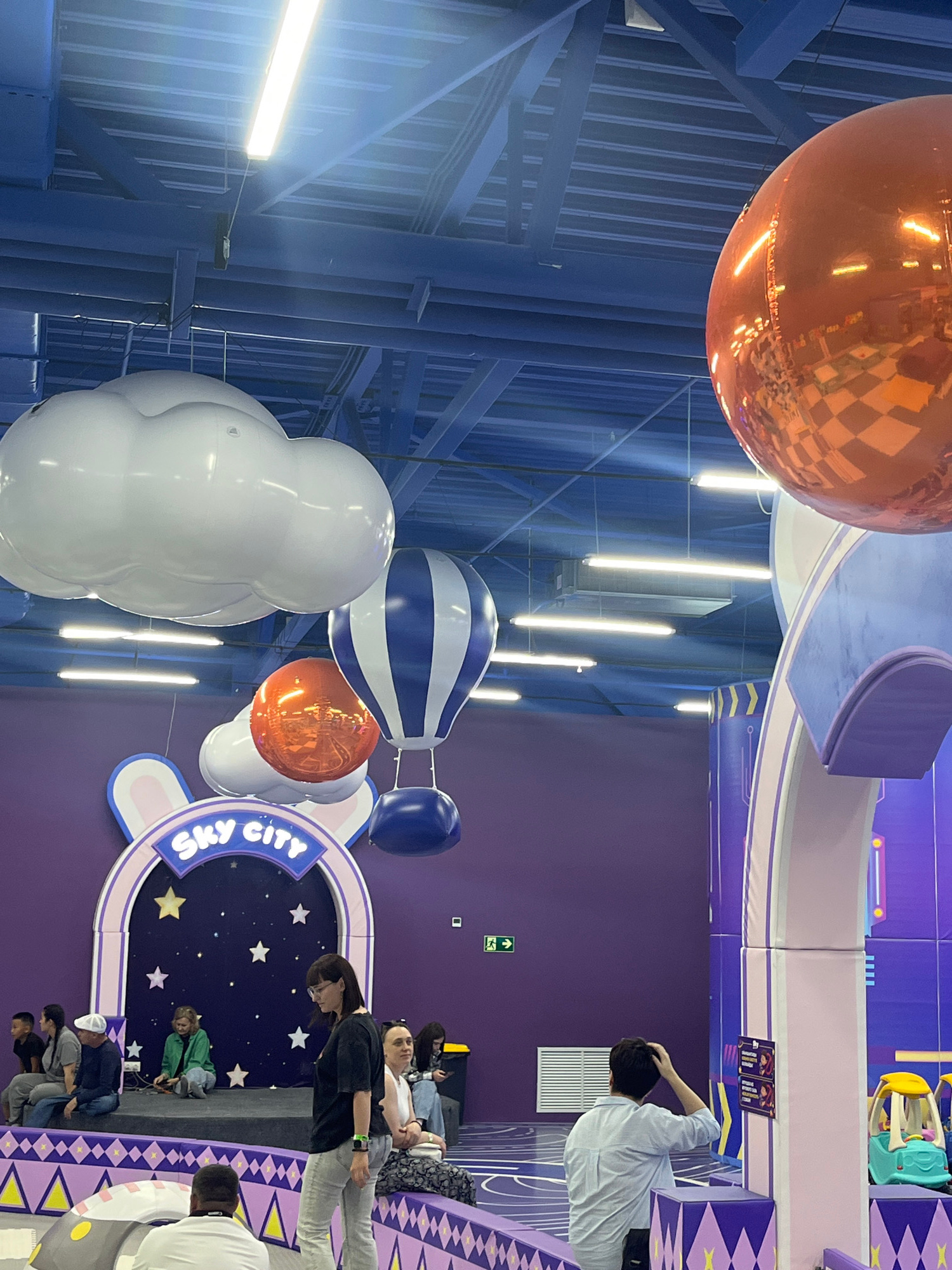 The image depicts the interior of "Sky City," an indoor entertainment center located in Kostanay, Kazakhstan. The scene features a vibrant, colorful environment designed for children. Prominently displayed are large, inflatable hot air balloons and clouds suspended from the ceiling, along with a large, reflective orange sphere. The walls are painted in shades of purple and adorned with playful graphics, including stars and a rabbit-themed archway above a stage area. Several people, including adults and children, are present. Some are seated, while others are standing and interacting. In the foreground, a person is seen from behind, with their hand on their head. In the background, there are play structures and a small, toy vehicle. The lighting appears to be from overhead fluorescent fixtures. The overall impression is that of a lively and engaging indoor playground.