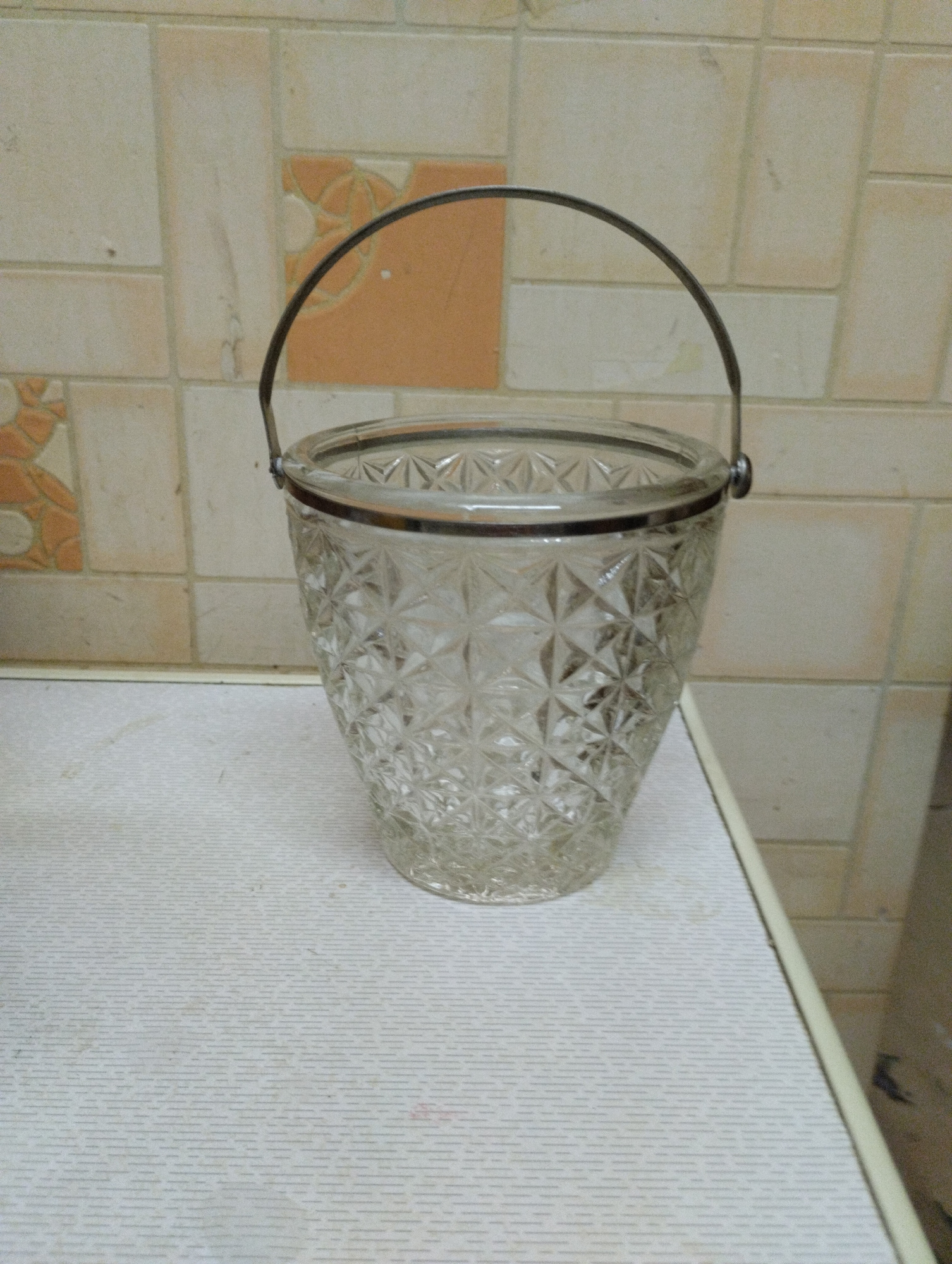An empty, clear glass ice bucket with a diamond-cut pattern sits on a white, textured surface. The ice bucket has a silver-colored metal handle arched over its top and a silver rim around the opening. The background consists of light-colored tiles, some of which are a pale orange with decorative patterns. The scene appears to be indoors, possibly in a kitchen or dining area. The image does not provide any information that would allow for the determination of its location in Tashkent, Uzbekistan.