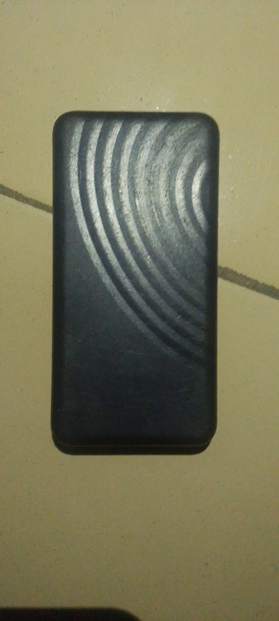 A black rectangular object with rounded corners and a textured, concentric circular pattern on its surface lies flat on a light-colored, textured surface. The object is positioned slightly off-center, towards the bottom left of the frame. The surface it rests on appears to be a tiled floor, possibly in an outdoor setting, given the faint diagonal lines that could represent grout or a shadow. The lighting is dim, casting subtle shadows around the object. There are no people or any other discernible objects in the immediate vicinity. The scene does not provide any specific visual cues to determine its location within Jauro Yinu, Nigeria.