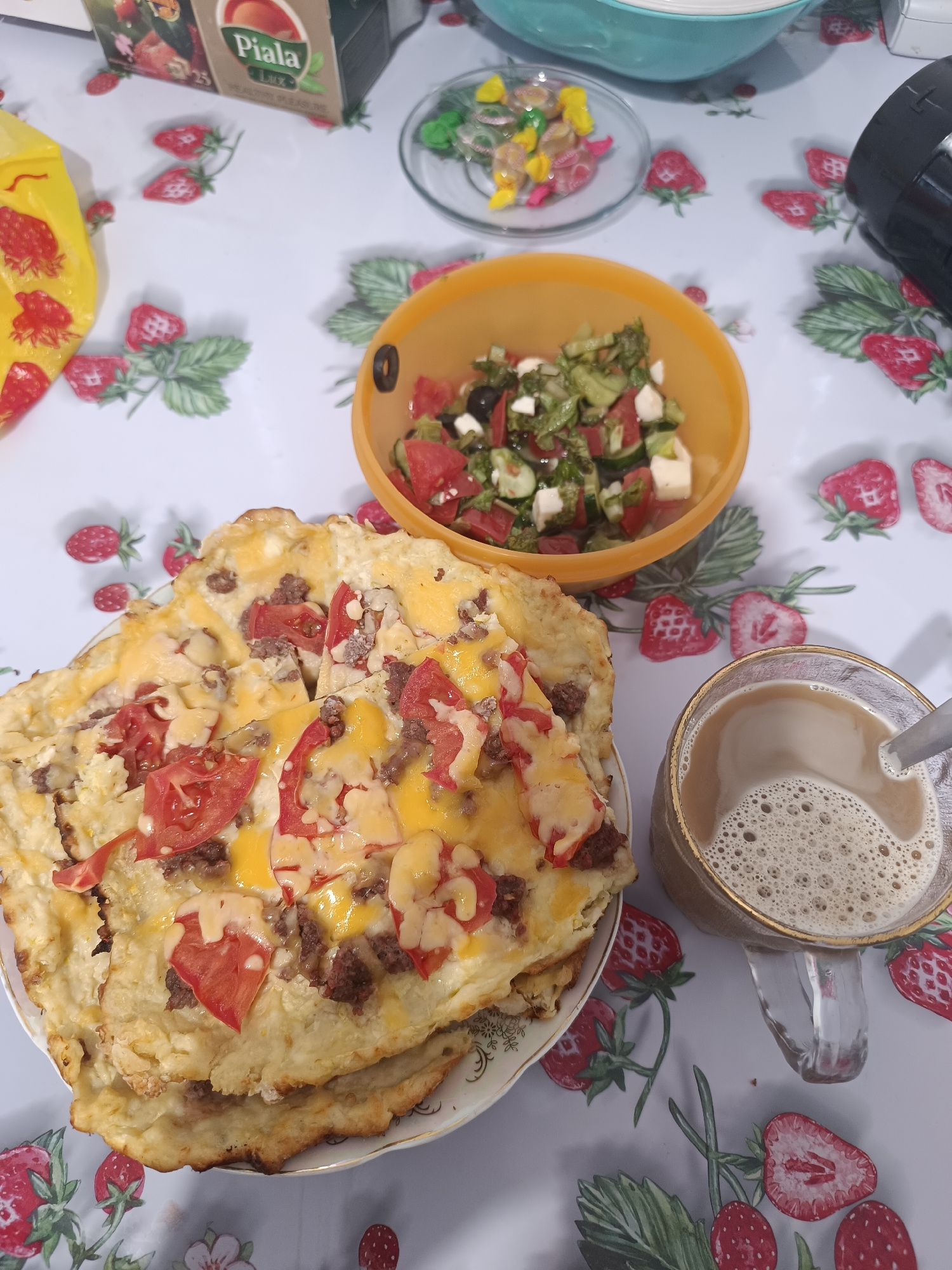 An overhead, medium shot captures a meal spread on a table covered with a white tablecloth adorned with a strawberry pattern. In the foreground, a clear glass mug filled with a frothy, light brown beverage, likely coffee or tea, sits to the right. A silver spoon is partially submerged in the drink. To the left of the mug, a plate holds a flatbread topped with melted cheese, tomato slices, and ground meat. A serving of salad, consisting of chopped tomatoes, cucumbers, olives, and white cheese cubes, is presented in an orange bowl behind the flatbread. A clear glass dish containing colorful candies is visible further back, near a box labeled "Piala." The background shows a portion of a blue bowl. The scene is indoors, on a table. There are no people present in the image. The location is not specified to be in Taraz city administration, Kazakhstan.
