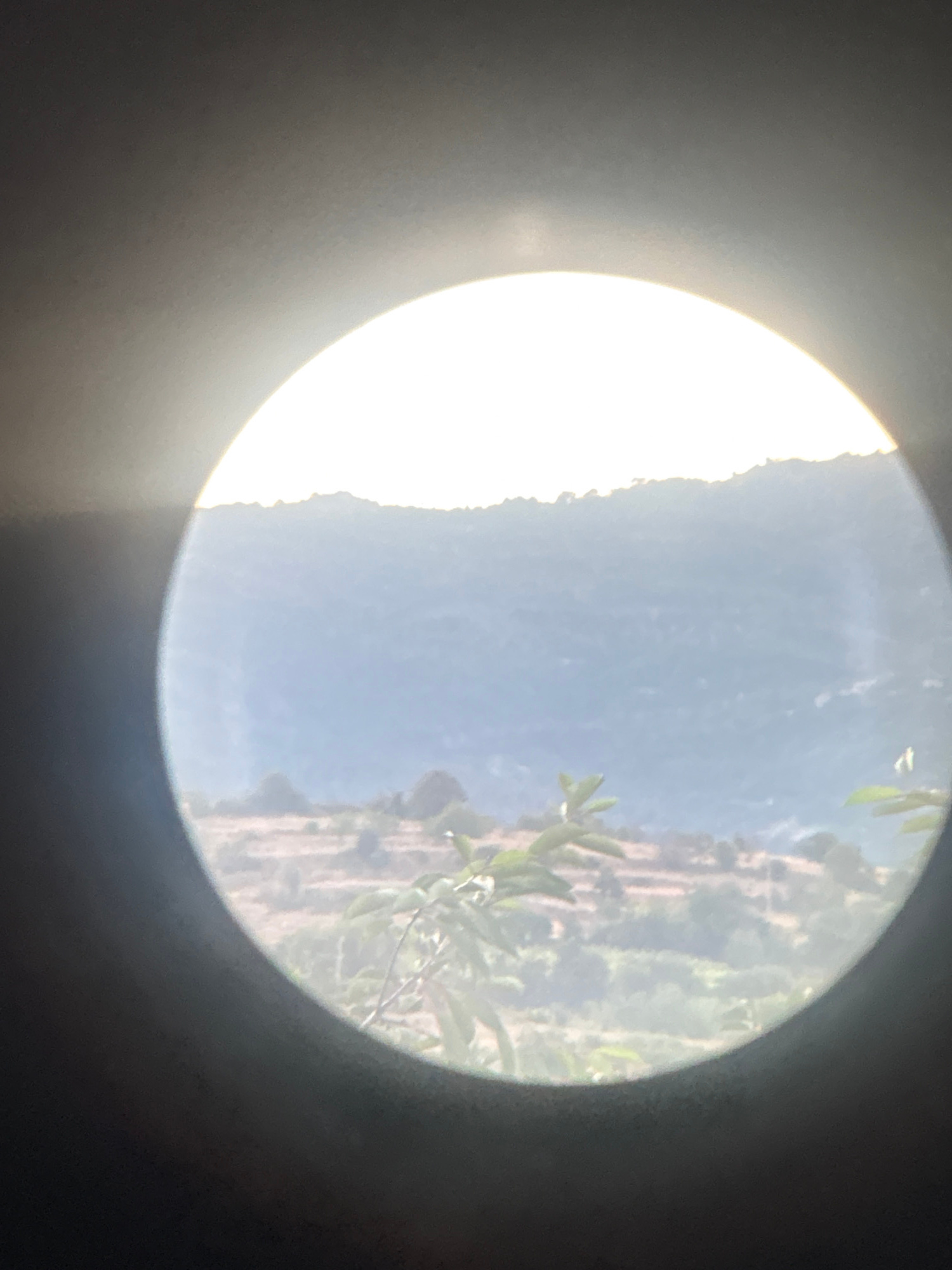 The image appears to be a circular view through a lens, possibly a telescope or camera lens. The view is of a natural landscape with rolling hills and vegetation. The sky is bright and appears to be either sunrise or sunset due to the diffused light. In the foreground, there are some green leaves, suggesting the viewpoint is from near some foliage. The background shows terraced hills with sparse vegetation and some structures that are difficult to discern clearly due to the distance and lighting. The scene is somewhat hazy, which could be due to atmospheric conditions. The location is described as Strelcha, Bulgaria.