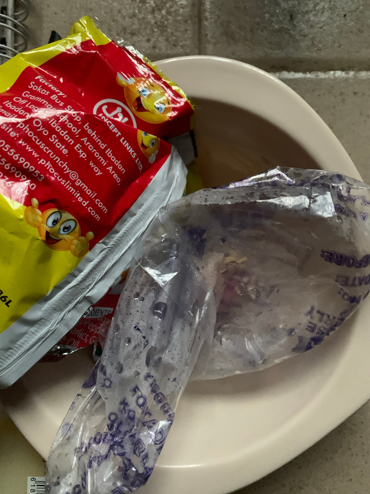 A close-up, eye-level shot shows a light-colored ceramic bowl containing several items. In the foreground, a clear plastic bag, slightly wet and with condensation, holds what appears to be a white substance. To the left of the plastic bag is a red and yellow foil package, partially obscuring it. Text is visible on the red portion of the foil, including "Factory:", "Sokas Bus Stop, behind Ibadan Grammar School, Araromi Area, Ibadan Oyo State", and a website address "www.phocuslimited.com". Below this, the word "INGREDIENTS" is visible, followed by "Pepper, Garlic, Oil". The background is a textured, gray surface, likely a wall. The lighting is moderate, creating some shadows and reflections. The scene appears to be indoors.