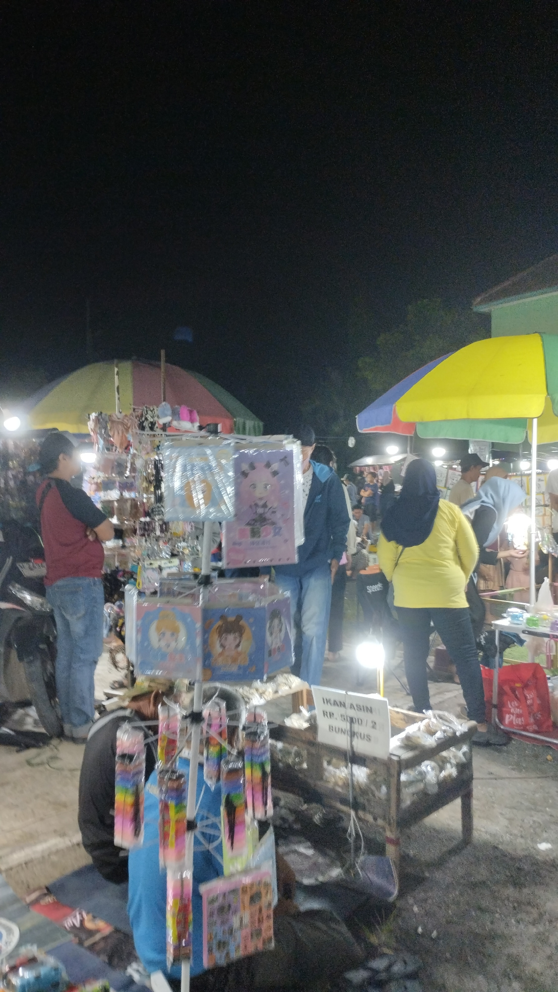 The image captures a nighttime outdoor market scene in Sambungrejo, Indonesia. A stall displays various items, including colorful accessories like hair clips and hair ties packaged in plastic. In front of the stall, a young person wearing a blue shirt and black pants sits on the ground, partially obscured by the display. To the right, a wooden table is laden with packaged dried fish, with a sign above reading "IKAN ASIN RP. 5.000 / 2 BUNGKUS". Several individuals are browsing or working at the market, some standing and others walking, under the illumination of artificial lights. The background features market tents and trees, suggesting a bustling local gathering.
