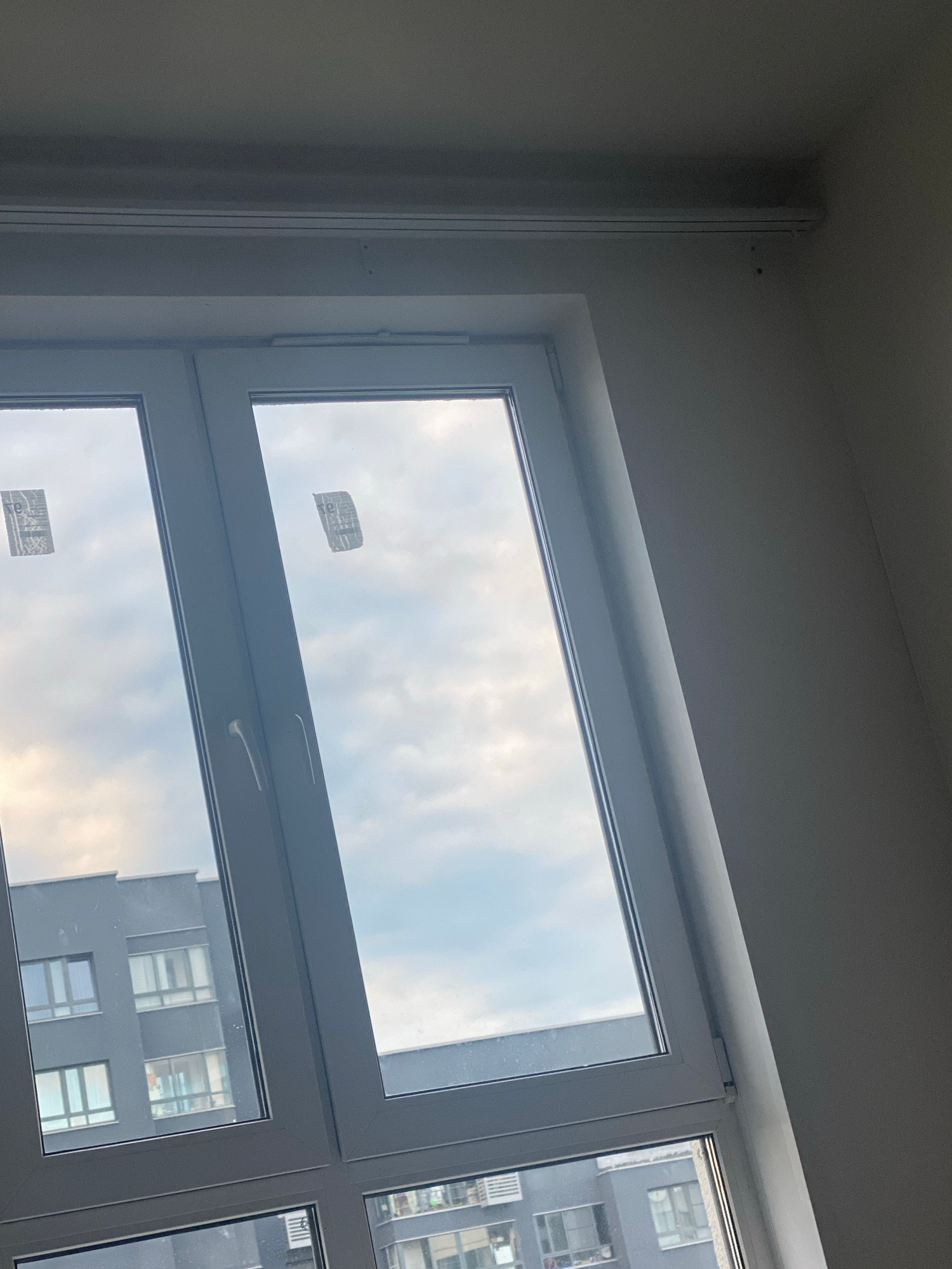 A wide-angle, slightly tilted exterior shot captures a modern, white-framed window against a neutral wall. The window is divided into two vertical panes, with a slight gap between them. Both panes reflect a cloudy sky, with hints of buildings visible in the reflection of the left pane, specifically a dark grey apartment building with multiple windows. Two rectangular stickers, appearing to be warning labels or product tags, are affixed to the inside of the window panes. The top edge of the frame reveals a simple white trim, and a thin, horizontal light fixture is mounted above it. The overall scene suggests an interior view looking outwards, possibly from a newly constructed building or an apartment.