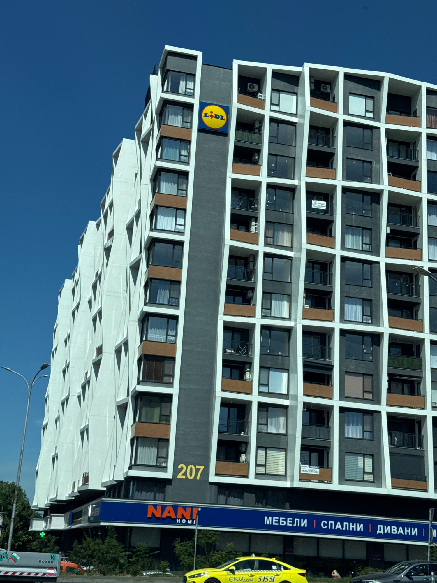 The image shows a modern residential building with a unique, angular white facade on the left and a dark gray facade with a grid of windows and balconies on the right. A prominent yellow Lidl logo is visible on the dark gray section of the building. The number "207" is displayed in large yellow numerals. At the base of the building, a blue sign with white lettering reads "NANI HOME" and "МЕБЕЛИ | СПАЛНИ | ДИВАНИ |" which translates to "FURNITURE | BEDROOMS | SOFAS |". A street with trees and a traffic light can be seen in the foreground. A yellow taxi cab with the number "51551" is parked on the street, and other vehicles are present. The sky is a clear, bright blue. The architectural style and the presence of the Lidl store and Nani Home furniture store suggest this is an urban commercial and residential area, likely in Burgas, Bulgaria, given the context provided.