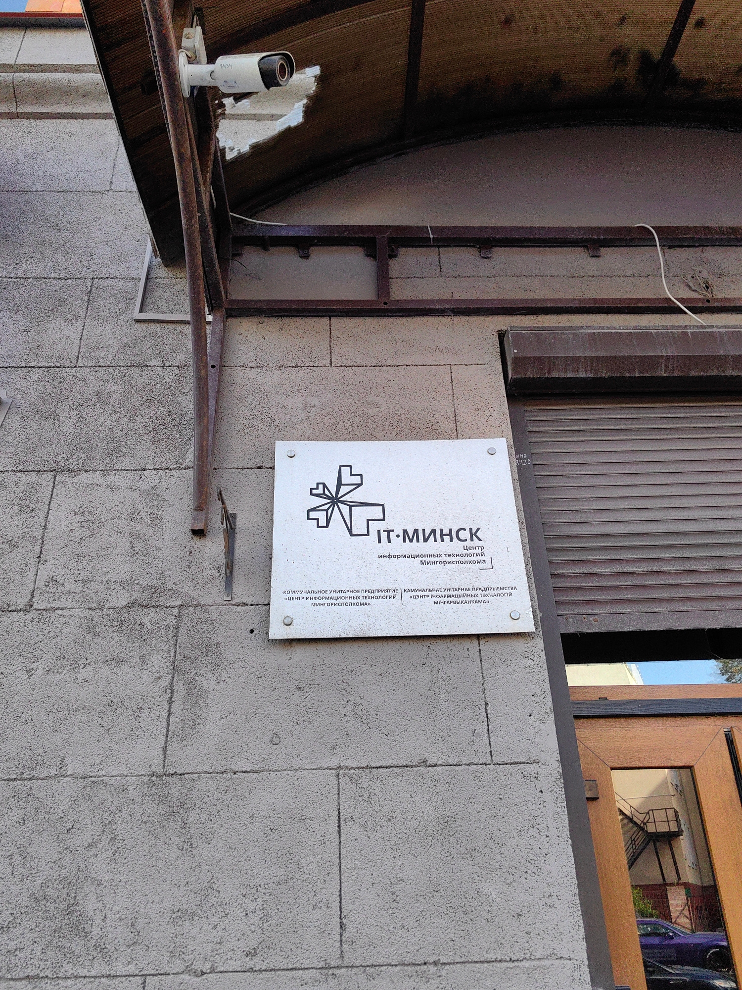 A security camera is mounted on a wall, attached to a rusty metal bracket under a brown awning. A white metal sign with black text and a stylized graphic is also affixed to the wall. The sign reads "IT-MINSK" in large letters, followed by smaller text that appears to be a translation or more detailed information. The wall is constructed of large, rough-textured concrete blocks. To the right of the sign is a closed, dark-colored rolling shutter and a wooden-framed doorway. The scene is outdoors, with daylight visible, and suggests an urban environment. The text on the sign likely indicates this is an IT-related facility in Minsk, Belarus.