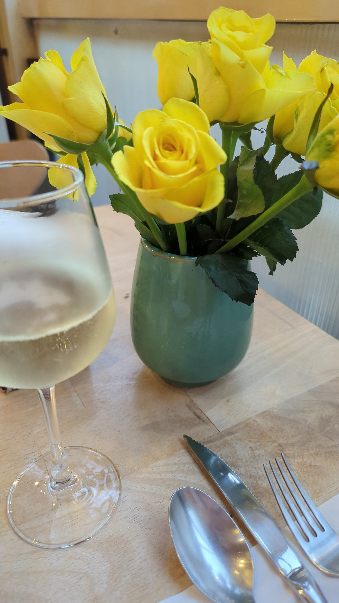 A close-up, slightly angled shot shows a table setting with yellow roses and a glass of white wine. In the foreground, to the left, is a wine glass filled with a light-colored liquid, possibly white wine, with visible bubbles. The glass has a clear stem and base. To the right of the glass is a small, teal-colored ceramic vase holding a bouquet of yellow roses. Several roses are in focus, with their petals open and vibrant. Their green stems and leaves are also visible. Further to the right, and slightly out of focus, are silverware: a spoon, a knife, and a fork. The table surface appears to be made of light-colored wood with a visible grain. The background is blurred, suggesting an indoor setting, possibly a restaurant or cafe. There are no people visible in the image. The overall impression is one of a still life arrangement, likely set for a meal or a decorative element.
