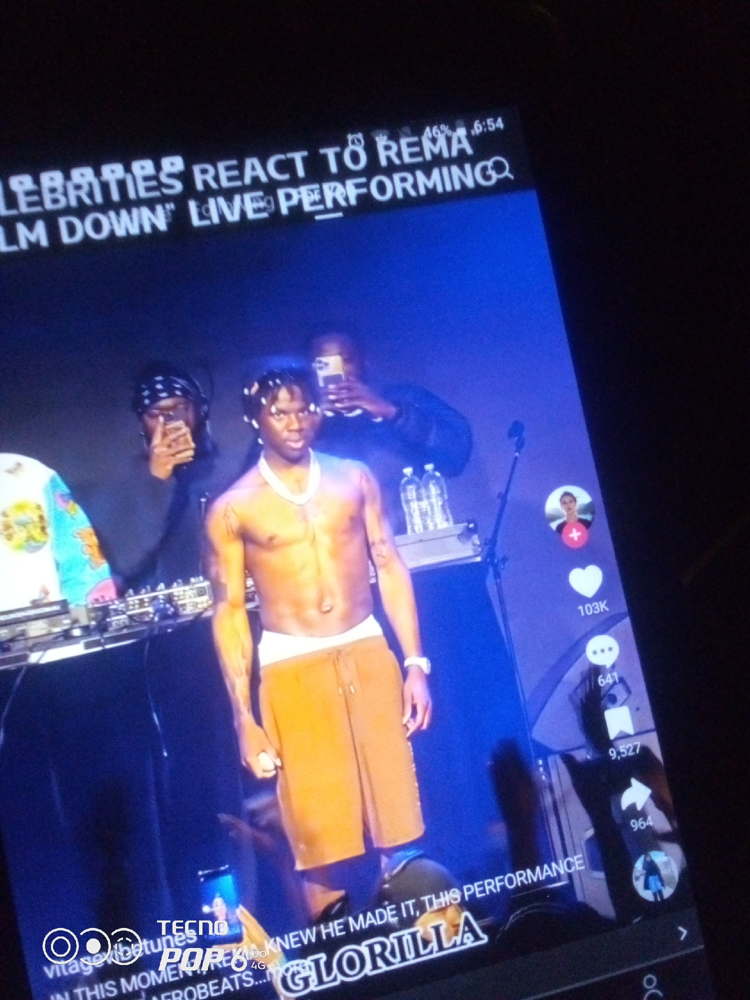 The image captures a screen displaying a video, likely from a social media platform, featuring a live musical performance. At the top of the screen, a title reads "CELEBRITIES REACT TO REMA 'CALM DOWN' LIVE PERFORMING." The main subject in the video is a shirtless male performer with braided hair, wearing a white necklace and orange shorts, standing on a stage. To his left, a DJ is visible behind a console, and another person wearing a bandana and glasses holds up a phone. Behind the performer, several bottles of water are stacked on a shelf. On the right side of the screen, social media interaction icons are present, including a profile picture with a "+" symbol, a heart icon with "103K" below it, a comment icon with "641" below it, and a share icon with "964" below it. At the bottom of the screen, text appears to read "IN THIS MOMENT FROBEATS... GLORILLA." There is no indication of the specific location within Howayi, Nigeria, being depicted.