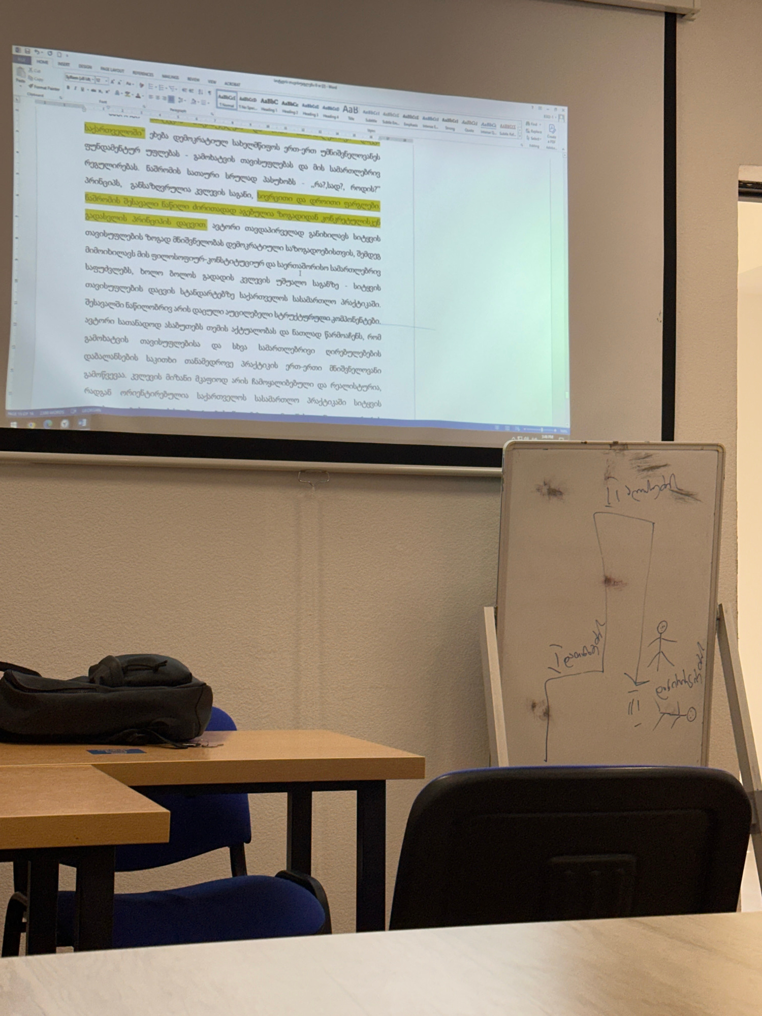 The image shows a classroom setting with a projector screen displaying a document in Georgian. The document is a text with highlighted sections. To the right of the screen is a whiteboard with some handwritten notes and a simple diagram. In the foreground, there are two wooden tables and two blue chairs. A black backpack is placed on one of the tables. The overall lighting suggests the room is indoors during the day, possibly with artificial lighting. The scene appears to be a typical university lecture hall or meeting room. Without further information or visual cues, it is impossible to determine the specific location within the city of Tbilisi, Georgia.