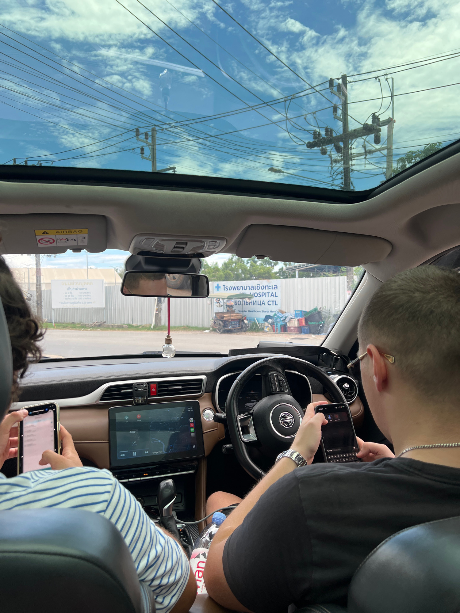 Two individuals are seated inside a car, with the camera positioned from the back seat. The driver, a male with short blonde hair, is wearing a black t-shirt and a silver chain, and is looking at and operating a smartphone. The passenger in the front seat, partially visible from behind, is wearing a blue and white striped shirt and is also holding and looking at a smartphone. The car's interior features a brown dashboard, a large touchscreen display showing a map, a steering wheel with the MG logo, and a panoramic sunroof revealing a bright blue sky with clouds and numerous power lines overhead. Outside the car, a sign in Thai and English, indicating "HOSPITAL CTL," is visible, along with construction materials and a motorbike. The scene appears to be in an urban or semi-urban area in Choeng Thale, Thailand, given the signage and infrastructure.