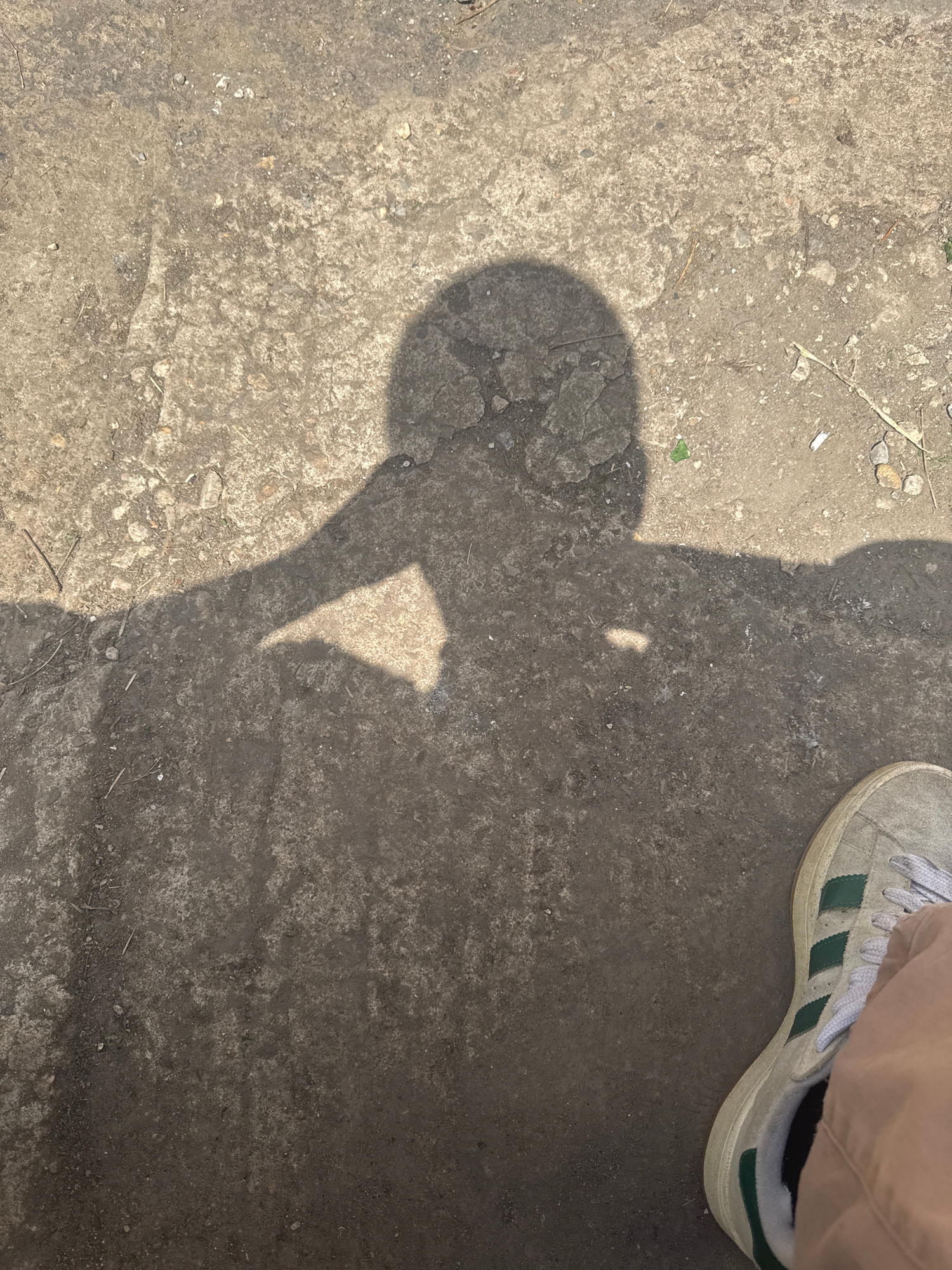 The image captures a shadow of a person's head and arm extended across a textured, grey concrete surface, suggesting an outdoor urban environment in Sofia, Bulgaria. The shadow, cast by sunlight from the left, shows the person's head in profile, with their arm raised and hand near their forehead, possibly indicating a gesture of thought or shielding from the sun. The concrete ground is uneven, with scattered small pebbles, dirt, and what appear to be dried grass or twigs. In the lower right corner of the frame, a portion of a light-colored sneaker with green stripes and white laces is visible, along with part of a person's leg clad in light brown trousers, confirming the presence of a person in the scene. The overall impression is that of a candid, everyday moment on a city street or pathway.