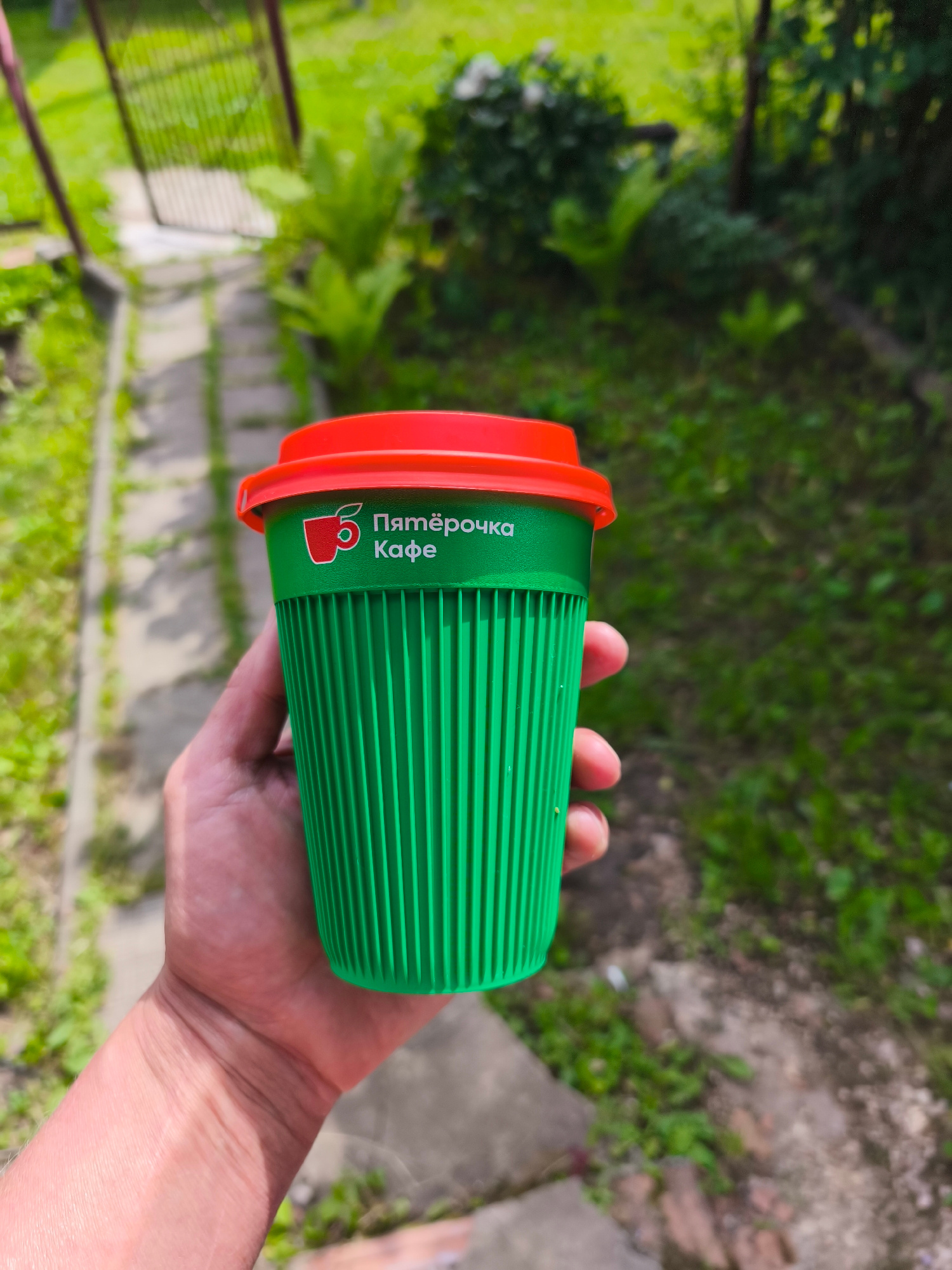 A person's hand holds a green disposable coffee cup with a red lid. The cup has a ribbed texture and features a white logo with the text "5 Пятёрочка Кафе" in Cyrillic script. The background is a slightly blurred outdoor scene, suggesting a natural setting with green foliage and a pathway. The lighting indicates it is daytime, likely sunny. The image seems to have been taken in a casual outdoor environment, possibly a backyard or garden, given the presence of plants and what appears to be a fence or gate in the upper left corner. The focus is on the coffee cup and the hand holding it, with the background providing context for the activity of enjoying a drink outdoors.