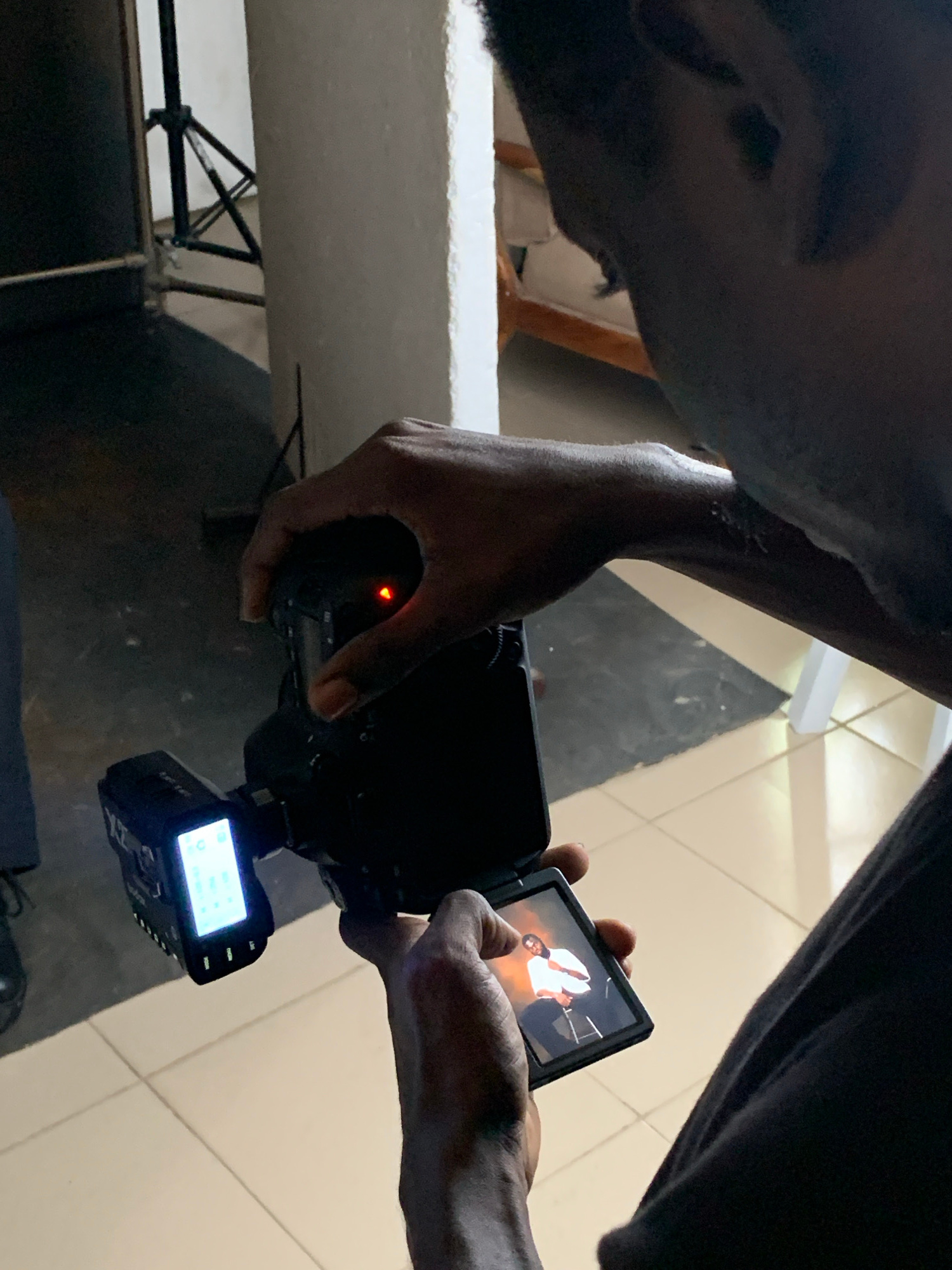 The image shows two individuals engaged in what appears to be a video recording session. One person, whose face is partially visible in the foreground, is looking down at a camera held by the other individual. This second person, with their hands holding a camera, is focused on the camera's screen. The screen displays an image of a person wearing a white shirt.
The setting is indoors, with a tiled floor and what looks like a white surface or desk in the background. There's also some lighting equipment visible. The time of day is not discernible from the image, nor is the weather. The overall impression is of a candid moment during a creative or professional activity. The individuals appear focused on their task, suggesting a deliberate and engaged process. No text is visible in the image. The location context of Jos, Nigeria is provided but not evident from visual cues within the image itself.