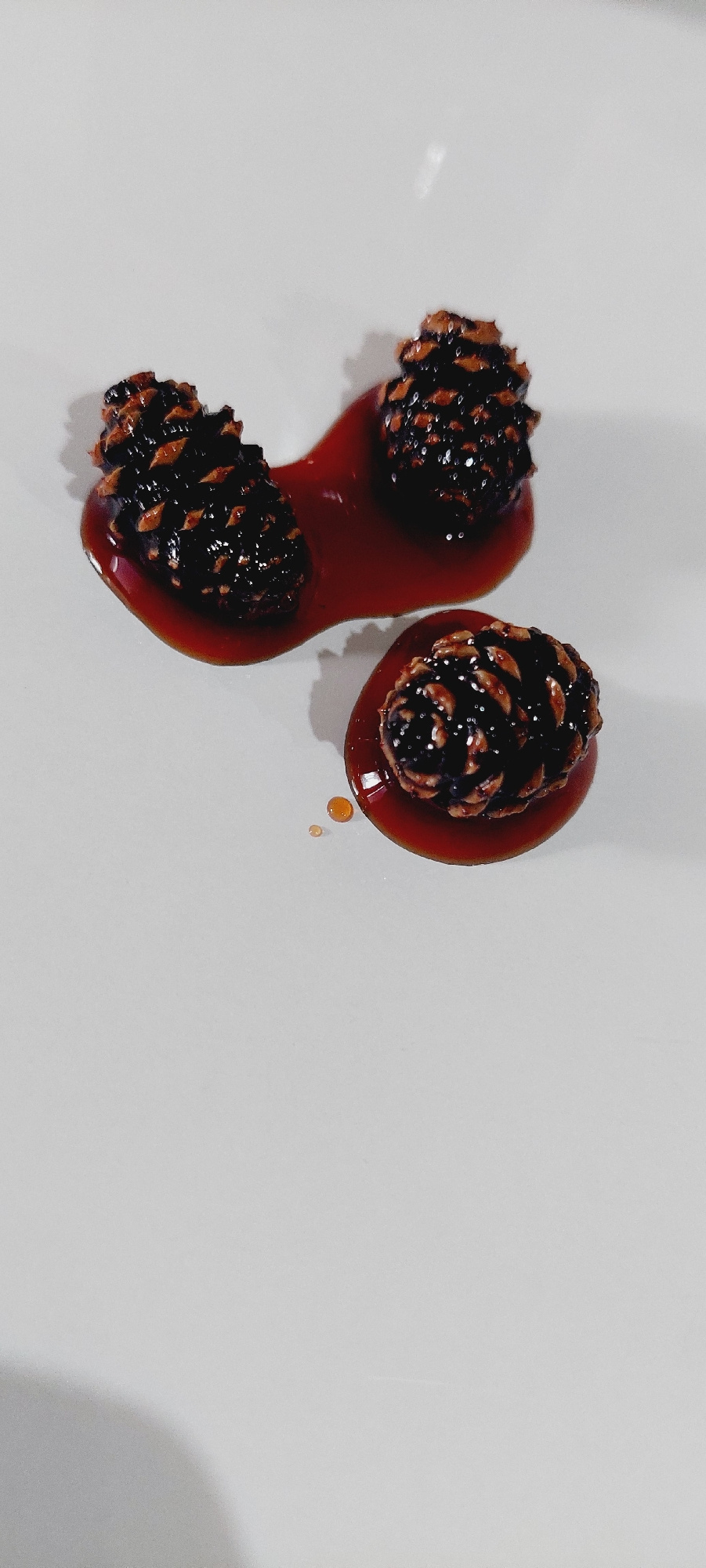 Three pine cones are arranged on a white surface. Two pine cones are positioned close together, partially submerged in a pool of reddish-brown liquid. A third pine cone is located below and to the right of the first two, also sitting in a small pool of the same liquid. Small droplets of the liquid are scattered on the surface near the pine cones. The lighting creates shadows on the white surface. The image does not contain any information or visual cues that would allow for its location to be identified in the city of Karachayevsk, Russia.