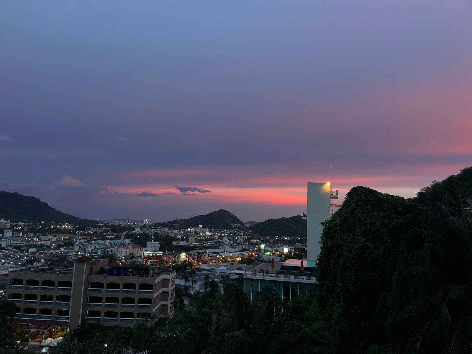 The image depicts a twilight cityscape, likely from an elevated vantage point overlooking the city of Ratsada, Thailand. The sky is a gradient of deep purple and pink hues, suggesting a sunset or pre-dawn twilight. The horizon is dominated by rolling, dark green hills, partially obscuring the city lights that dot the landscape below.
In the foreground, several buildings are visible. To the left, a multi-story building with a beige facade and numerous square windows is prominent. Its lower levels appear to have a covered entrance with parked cars. Adjacent to this is a smaller building with a reddish-brown tiled roof. To the right, a stark white, tall rectangular structure with an external staircase stands out, partially covered by dense, dark green foliage that also occupies the right edge of the frame, suggesting a thick natural growth. In the mid-ground, more urban structures are visible, including low-rise buildings with illuminated windows and signs, one of which reads "MATRIX." The overall impression is of a dense urban area nestled amongst natural hills under a dramatic sky. No people are discernible in the image.