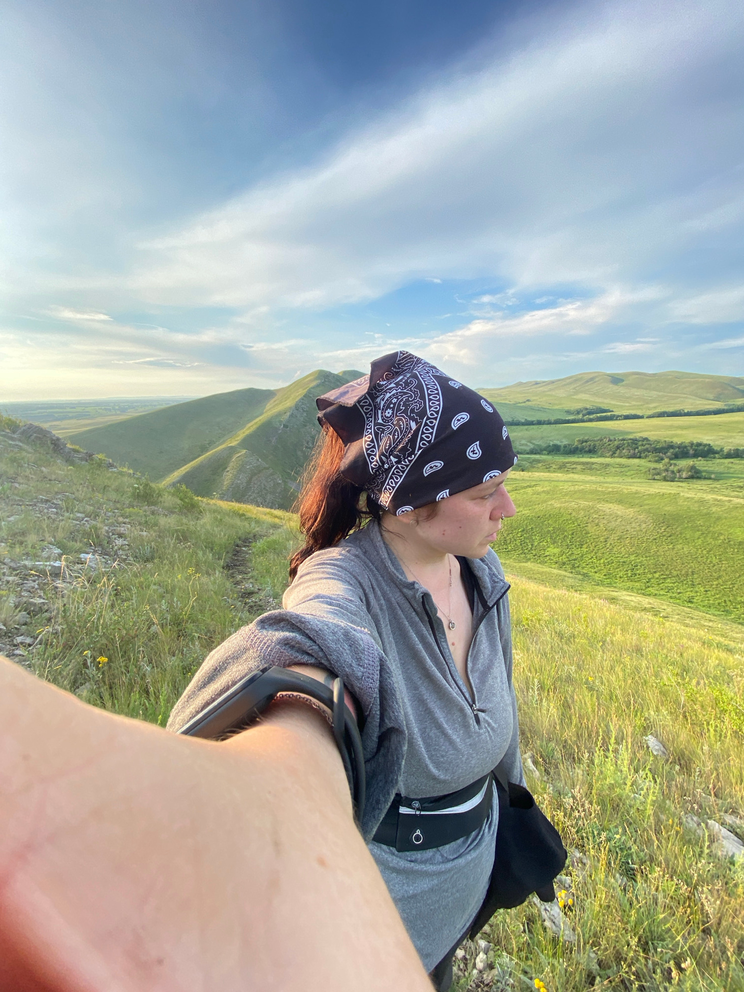 A selfie is taken by a person wearing a gray zip-up hoodie, a black bandana with a white paisley pattern, and a black fanny pack, on a grassy hillside. The person is looking to the right, with their left arm extended towards the camera, capturing a portion of their arm and hand. The background features rolling green hills and a bright blue sky with wispy white clouds, indicative of a sunny day. The location is identified as near Novogafarovo, Russia. The overall scene suggests an outdoor excursion or hike in a rural, hilly landscape during daylight hours.