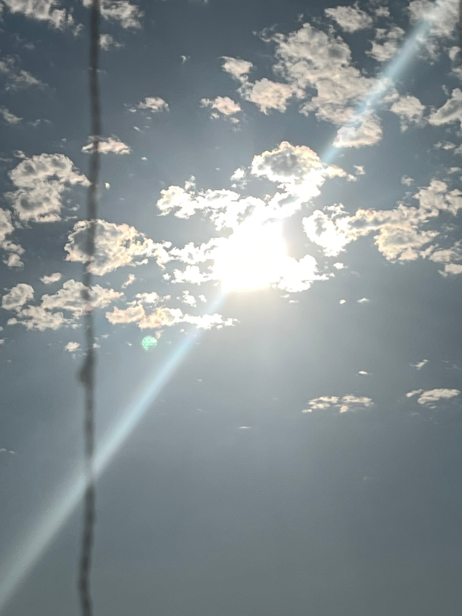 This media file captures a bright, sunny sky with scattered clouds, viewed from a low angle, possibly near the ground. The sun is a prominent, overexposed element in the center-left of the frame, emitting a strong glare that casts a diagonal light ray across the image. This ray is partially diffused by some of the clouds, creating a hazy effect.
The clouds themselves are varied in size and shape, appearing white and fluffy against a greyish-blue sky. Some clouds are backlit by the sun, giving them a golden or yellowish hue, while others are in shadow. The overall impression of the sky is bright and potentially warm, suggesting daytime.
A thin, dark, horizontal line stretches across the lower portion of the image, appearing to be a wire or cable. This object is slightly out of focus, implying it is in the foreground.
There are no visible people or text in the image. The scene is purely natural, focusing on the sky and atmospheric conditions. The brightness of the sun and the clear, albeit cloudy, sky suggest fair weather. The image has a slight tilt, which adds a dynamic, almost abstract quality to the composition. Given the location context of Sochi, Russia, this could be a view of the sky over the city or its surrounding natural landscape. The time of day is clearly during daylight hours, and the presence of a strong sun indicates it is not overcast.