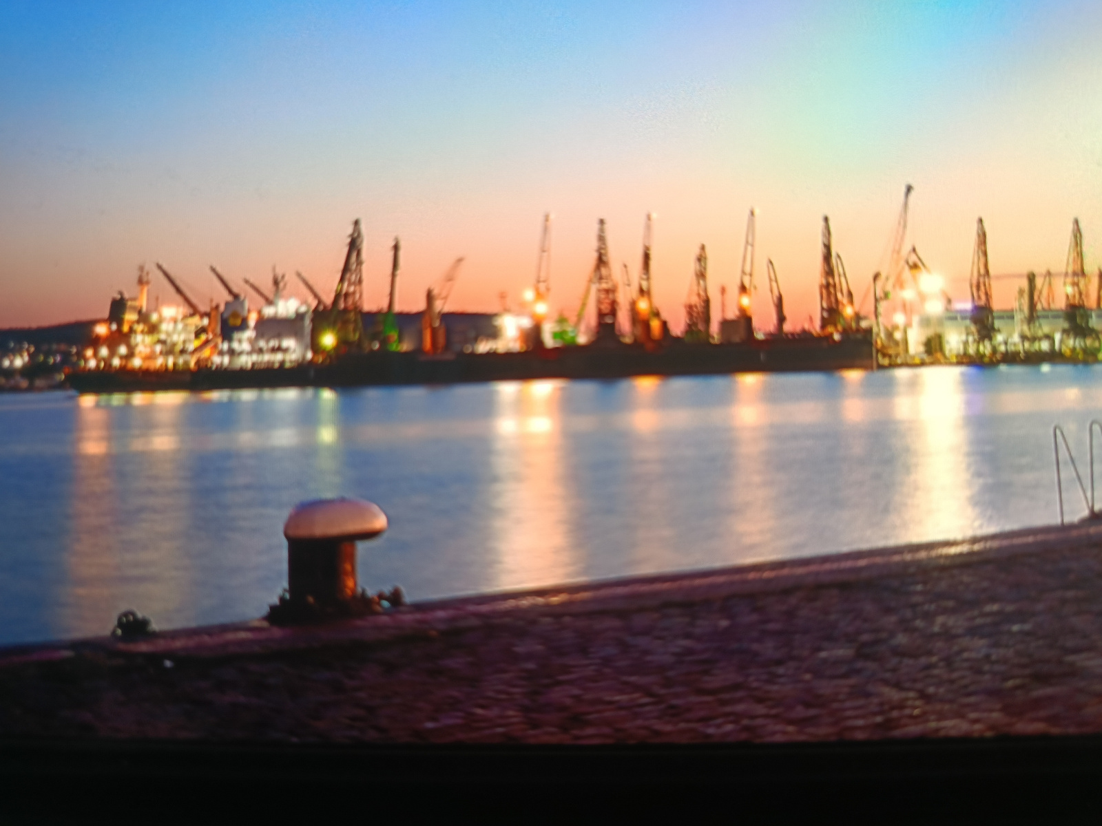 The image depicts a harbor scene at dusk, with a clear sky transitioning from blue to a gradient of orange and pink. The water in the foreground is calm, reflecting the lights of the industrial area on the opposite shore.
The industrial area is characterized by a line of large cranes, some of which are illuminated. Several ships are docked, and their lights also contribute to the overall illumination of the scene. The architecture of the industrial buildings is not clearly discernible due to the distance and lighting conditions.
In the immediate foreground, there is a dark, textured surface, likely a pier or embankment, with a prominent mushroom-shaped bollard. The bollard is dark with a light-colored cap. The surface of the pier appears uneven and possibly made of stone or concrete. A metal railing is visible on the far right of the pier.
The overall impression is one of a working harbor at the end of the day, with artificial lights creating a warm glow against the twilight sky. This scene is consistent with the industrial port facilities located in Varna, Bulgaria, specifically the Varna Port area which features numerous cranes and shipping infrastructure along the Black Sea coast. There are no people visible in the image.