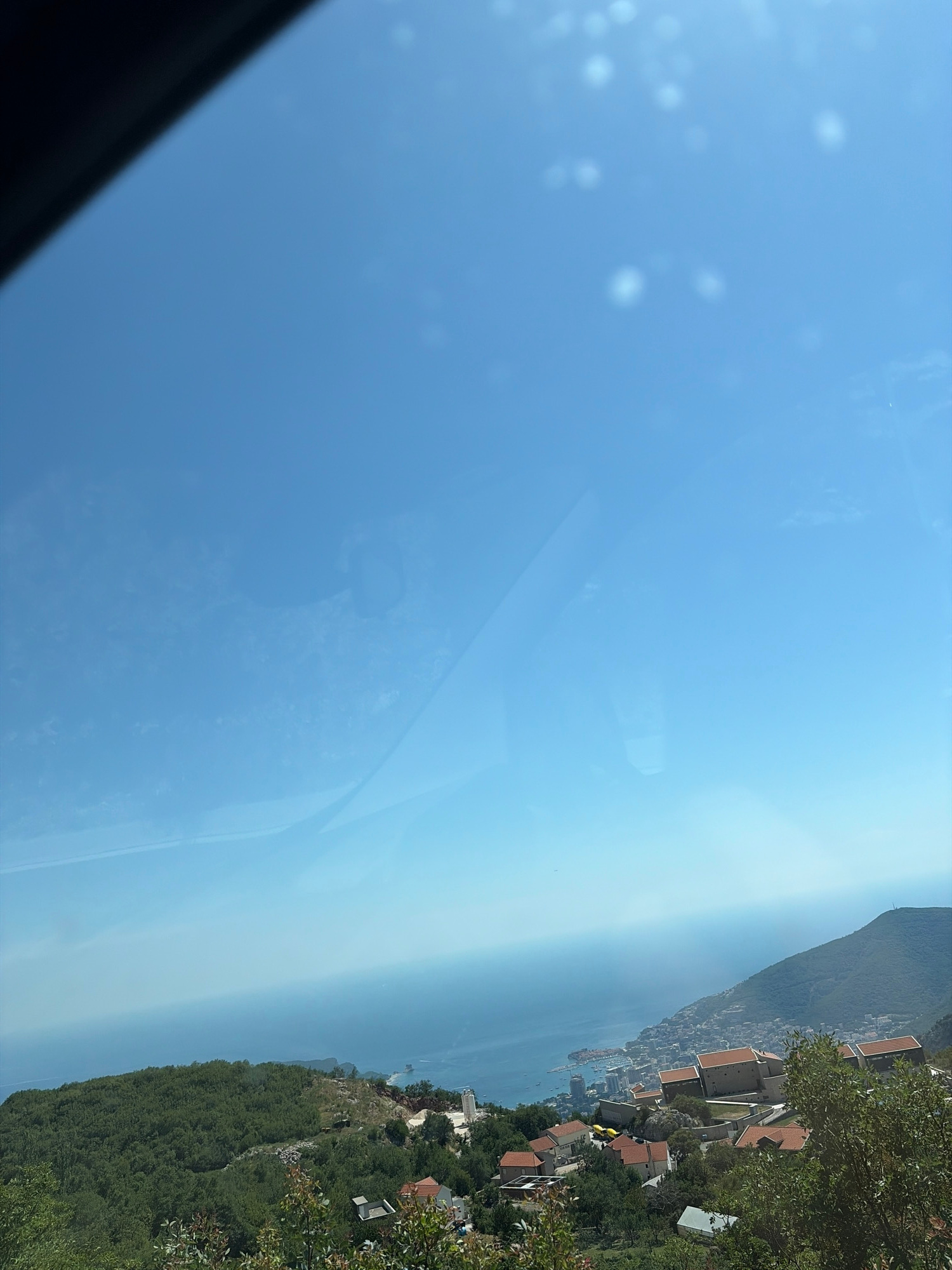 The media file displays a scenic view of a coastal town nestled against a mountainous backdrop. The scene is dominated by a vast expanse of blue sky, with some hazy atmospheric conditions softening the distant horizon where the sky meets the sea. On the right side of the image, a hillside is densely covered with lush green trees. Scattered amongst the foliage are several buildings with terracotta-colored roofs, suggesting a residential or resort area. Some of these buildings appear to be perched on the slope, offering views of the water.
The sea itself is a calm blue, stretching out towards the hazy distance. A coastline dotted with more buildings and infrastructure is visible along the water's edge, indicating a populated area. Boats can be seen on the water, hinting at maritime activity. The mountain range rises majestically behind the town, its slopes covered in vegetation, fading into a lighter blue haze in the distance. The overall impression is one of a bright, clear day, likely daytime, with a tranquil and picturesque setting. The sun is high in the sky, casting a soft light over the landscape. No people are explicitly visible, but the presence of buildings and boats implies human habitation and activity. The location cues, such as the style of architecture and the mountainous coastal terrain, are consistent with the provided location context of Lapčići, Montenegro. There is no visible text in the image.