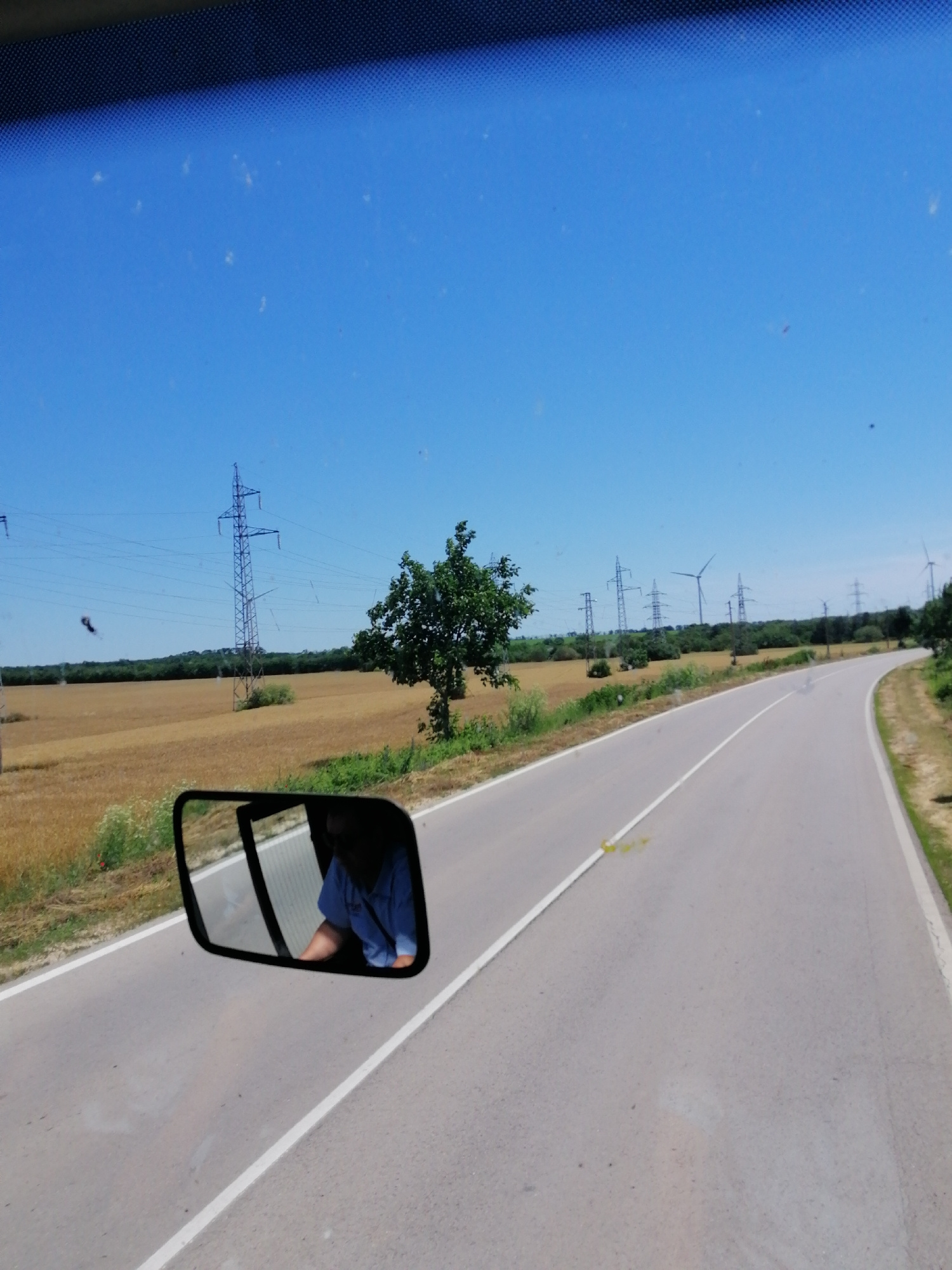 The image shows a view from inside a vehicle, looking out through the windshield. A side-view mirror on the left reflects a person wearing a blue shirt, seated inside the vehicle. The exterior scene features a two-lane asphalt road curving to the right. The surrounding environment is rural, with a field of golden crops on the left, bordered by trees and vegetation. Power lines and tall electrical pylons are visible in the background, along with several wind turbines. The sky is clear and blue. The location is described as Unknown, Unknown.