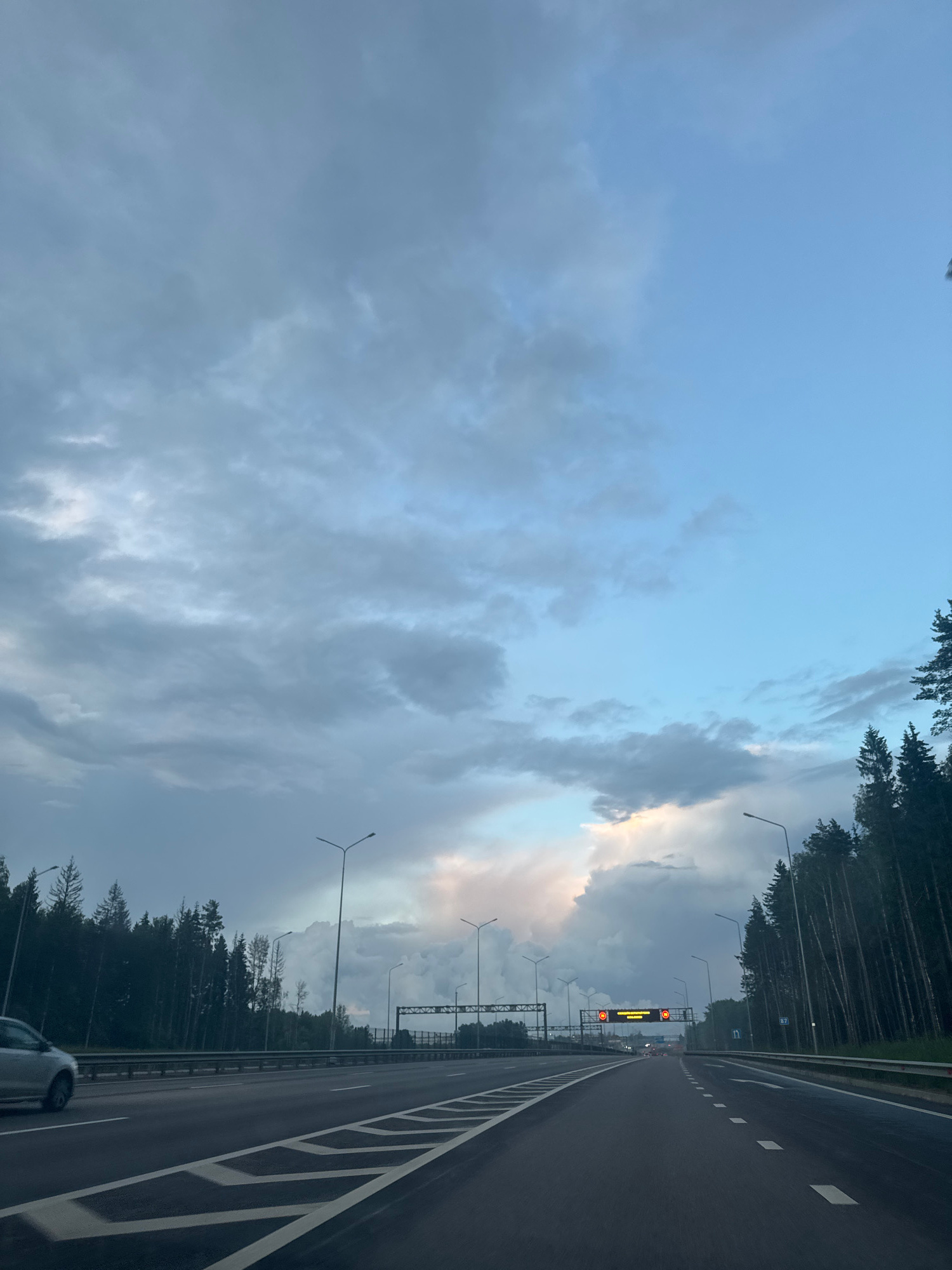 The image depicts a view of a highway with a dense forest bordering both sides. The sky is overcast with dramatic clouds, suggesting an impending storm or recent rainfall. There are streetlights lining the highway and overhead gantries with digital signs. The road itself is asphalt with white lane markings. A silver car is visible in the left lane, moving away from the camera. In the distance, there are additional overhead structures and trees. The location appears to be on a highway within or near the city of Shchyolkovo, Russia, given the context provided. The overall atmosphere is somber due to the cloudy weather and the imposing presence of the forest.