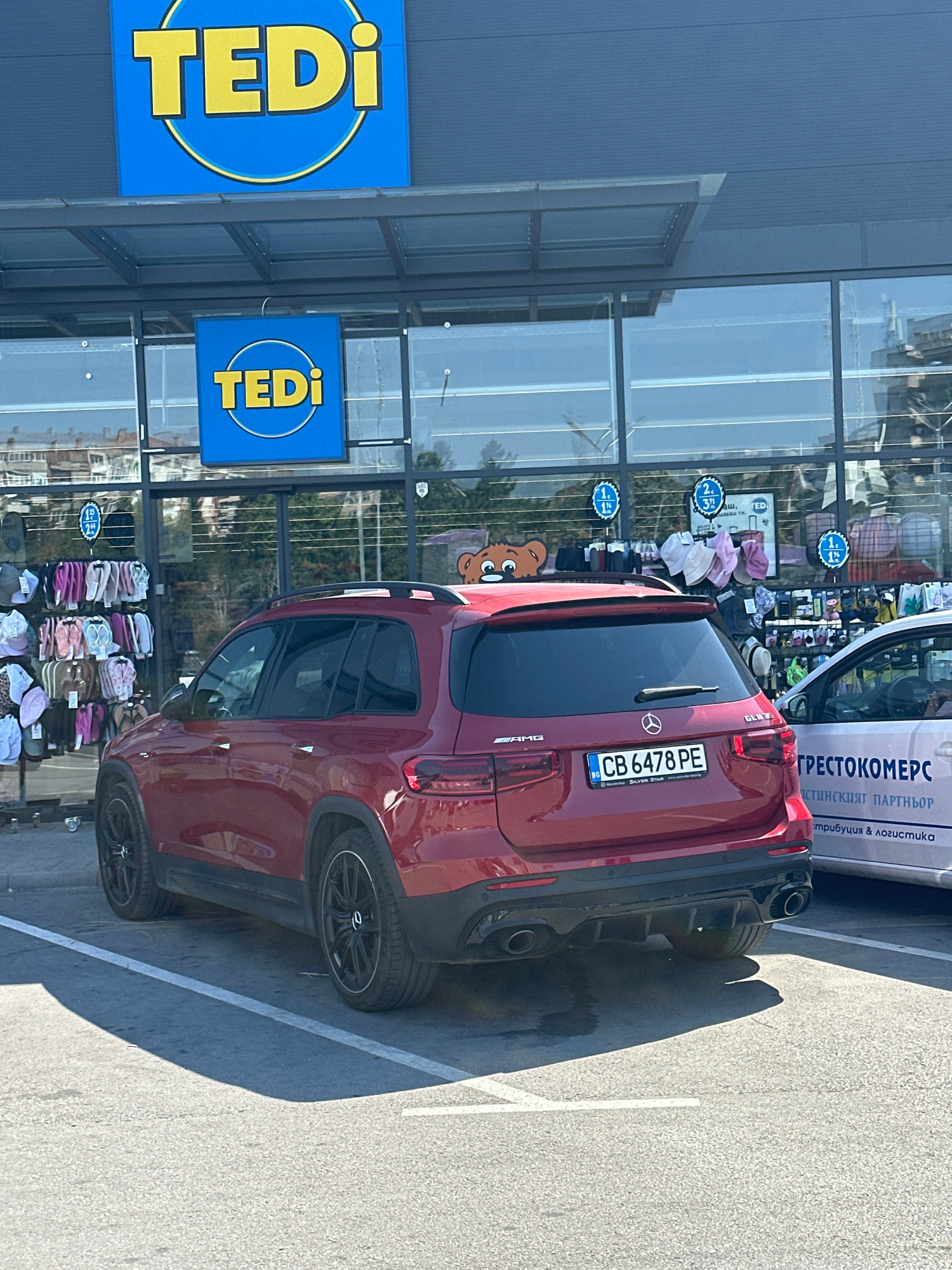 A deep red Mercedes-Benz GLB35 AMG is parked in front of a TEDi store. The car is facing away from the viewer, with its rear end prominently displayed. The TEDi logo, featuring a blue square with a yellow circle and the word "TEDi" in yellow, is visible on the store's facade. A smaller TEDi sign is also mounted on the glass windows.
Inside the store's windows, shelves are stocked with various items, including what appear to be hats and soft toys. The car has a Bulgarian license plate that reads "CB 6478 PE". The words "Silver Star" are visible below the license plate, and "AMG" is printed on the rear bumper. Text on a white vehicle parked to the right of the Mercedes reads "ГРЕСТОКОМЕРС" and "ИСТИНСКИЯТ ПАРТНЬОР", suggesting a business named "Gregostomers", a "true partner" specializing in "distribution & logistics".
The scene suggests daytime, with bright sunlight casting distinct shadows on the asphalt. The presence of the TEDi store indicates a commercial setting, likely a retail area. The overall mood is neutral, focusing on the objects and the environment.