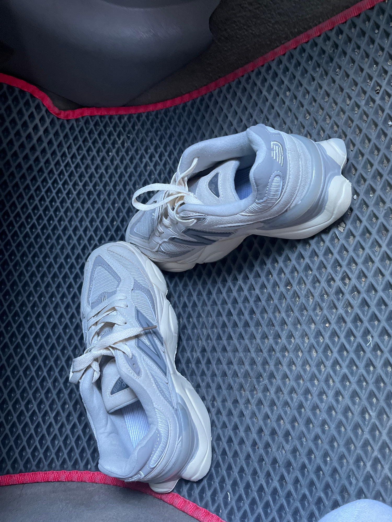Two white and gray athletic shoes are placed on a black, diamond-patterned mat. The mat has a red border on the top and bottom edges. The shoes are positioned diagonally, with the toe of one shoe pointing towards the upper right of the frame and the toe of the other shoe pointing towards the lower left. The lighting suggests an indoor setting, possibly a car interior or a covered outdoor area, given the texture of the mat and the dark background. There is no indication of the location being Ivanivka, Ukraine.