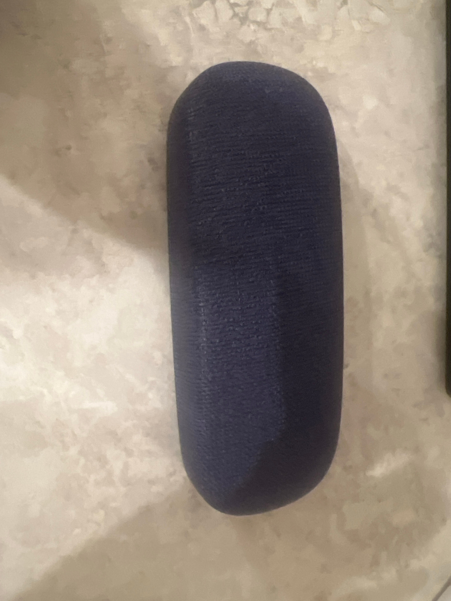 The image shows a dark blue, oblong glasses case resting on a light-colored, textured surface that appears to be tile or stone. The case is positioned vertically in the center of the frame and is slightly angled. The surface has a marbled or mottled pattern with shades of beige and off-white. There are no people or visible actions in the image. The lighting is somewhat dim, casting a subtle shadow on the left side of the case. The scene is indoors, and the location in Saratov, Russia, is not determinable from the visual information provided.