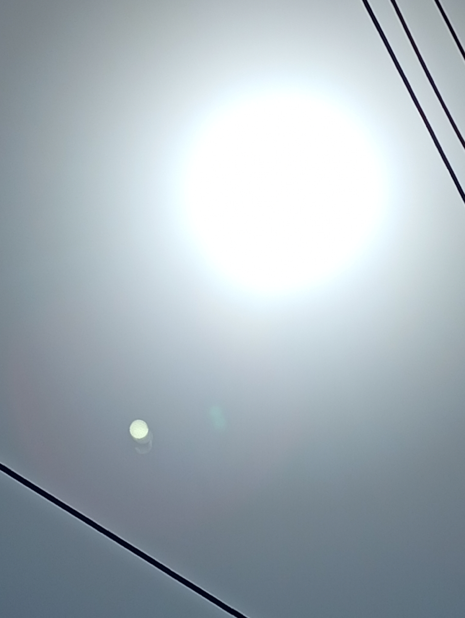 A bright, circular light source dominates the upper portion of the image, creating a lens flare effect that washes out the background. Below this, a smaller, circular light source is visible, possibly a reflection or another light fixture. Several dark, linear elements, likely cables or wires, extend diagonally across the frame, from the upper right to the lower left and upper left to the lower right. The overall environment appears to be indoors, with a neutral, gray-toned ceiling or surface as the backdrop.