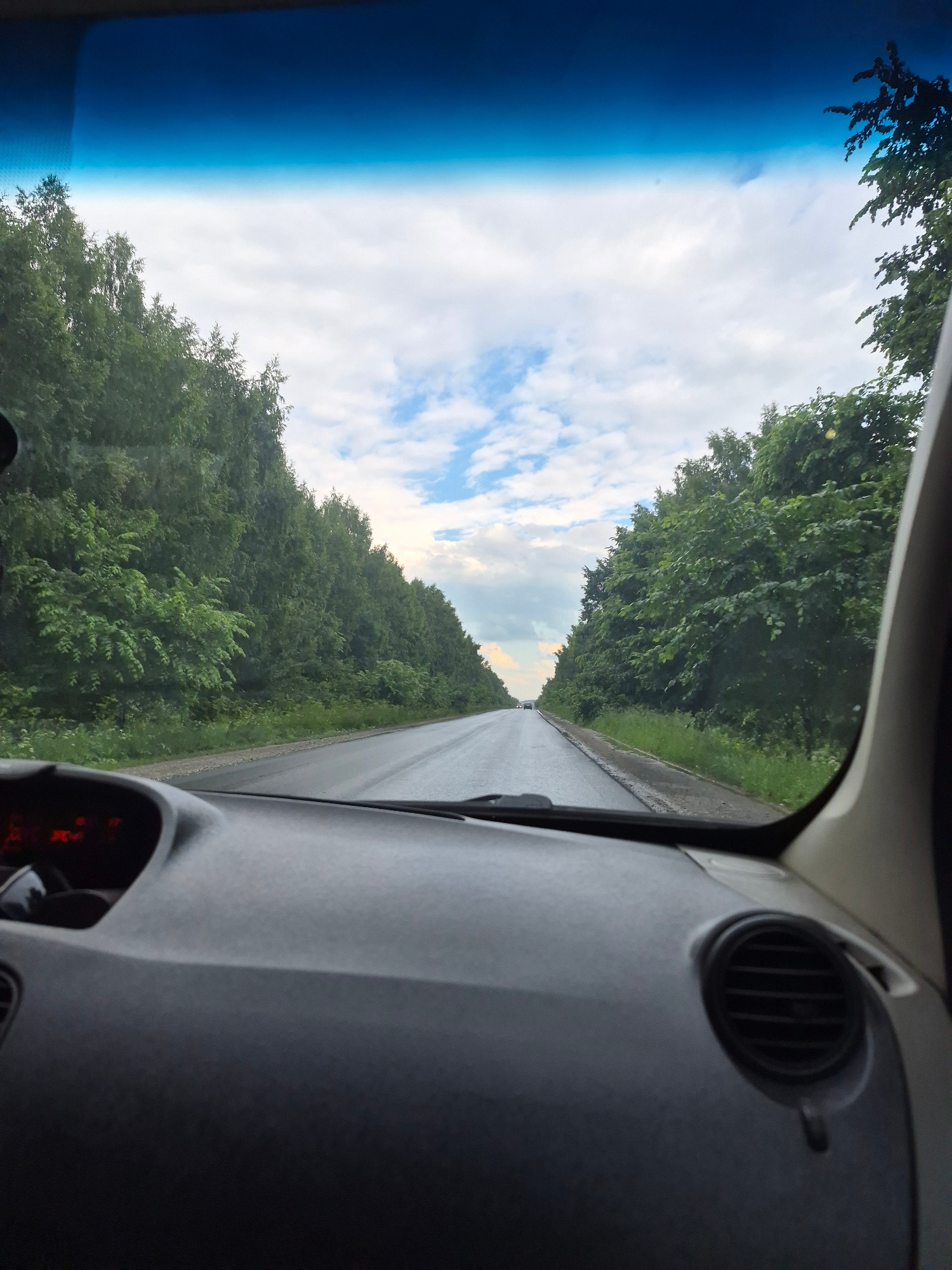 The image captures a view from inside a car, looking forward through the windshield. The scene depicts a paved road stretching into the distance, flanked by dense green trees on both sides. The sky above is partly cloudy, with patches of blue and white clouds. The sun appears to be low in the sky, casting a soft light and suggesting it might be early morning or late afternoon.
The interior of the car is partially visible, including the dashboard with an air vent and some red dashboard lights. The windshield has a blue tint at the top, likely from a sunstrip or tinting. The orientation of the image is vertical, and the perspective suggests the car is in motion on a rural road, possibly in a wooded area. The trees appear lush and full, indicating it is likely summertime.
There are no people visible in the image. The overall impression is one of a peaceful drive through a natural landscape. No text is visible in the image. Given the location context of Plamya-3, Russia, the scene likely represents a drive through the Russian countryside during the warmer months.