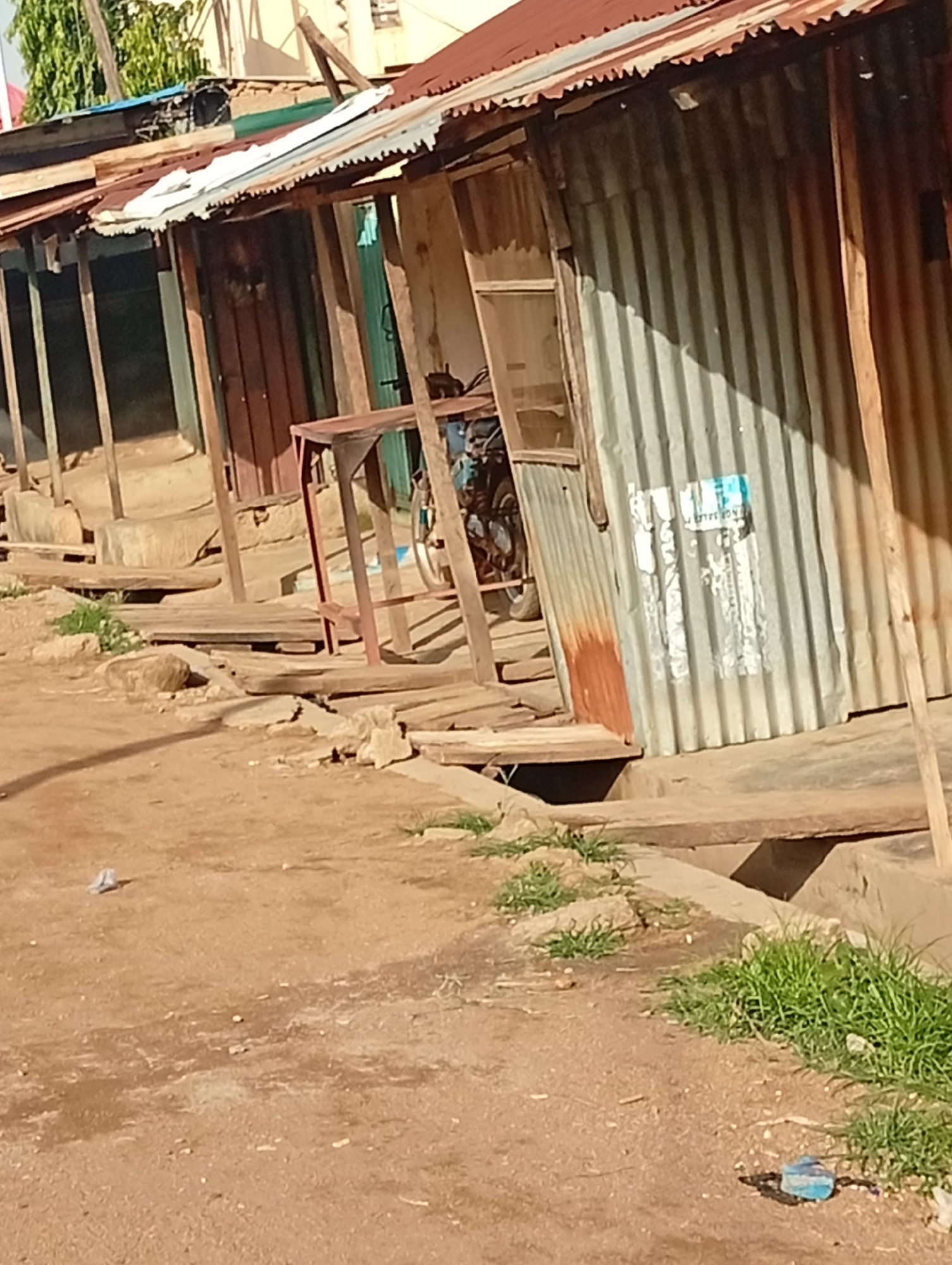 A low-angle, eye-level medium shot captures a weathered, corrugated metal structure in Sabon Tasha, Nigeria. The structure appears to be a shop or stall with a rusty, red metal roof supported by wooden beams. The walls are primarily corrugated metal, with some sections showing signs of rust and wear. A wooden plank walkway or platform runs in front of the structure. To the left, a portion of a reddish-brown wooden door is visible. A metal table with a reddish-brown frame stands in front of the structure. In the background, behind the metal walls, a light-colored building with a peaked roof and trees can be seen. The ground in the foreground is dirt, with scattered rocks, grass, and some pieces of litter, including a blue plastic bottle. The overall impression is one of a utilitarian, possibly informal, commercial space in an outdoor, possibly semi-rural or urban fringe, setting.
