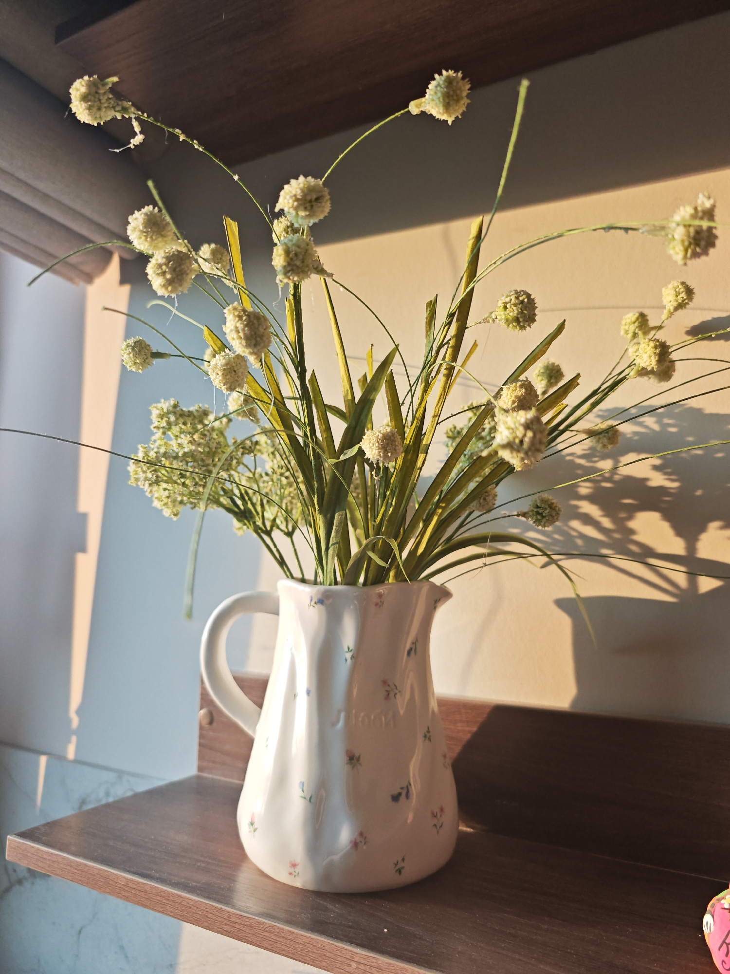 A still life composition features a white ceramic pitcher with small floral decorations, filled with artificial dried flowers and greenery. The pitcher is placed on a wooden shelf. Behind the pitcher, a wall is visible, casting shadows from the dried flowers. The scene is illuminated by sunlight coming from the left, creating strong highlights and shadows. The overall aesthetic is rustic and decorative. The image depicts an indoor setting, possibly a home or a shop, with no specific indicators pointing to Kazan, Russia.