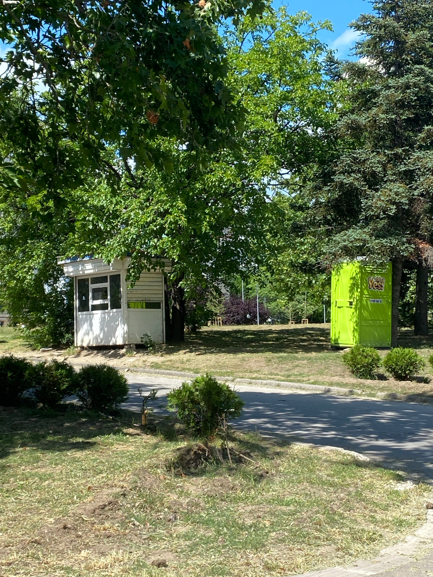 The image shows a scene with two structures amidst trees and greenery, located in Varna, Bulgaria. On the left, a small, white booth with windows and a blue roof is situated under the shade of a large tree. The booth appears to be made of a corrugated material with white siding and a window frame. To its right, a bright green portable toilet is positioned next to a tall evergreen tree. The portable toilet has a visible logo that says "YES!". The ground in front of the structures is covered with grass and some bare patches, with a paved area or road visible in the foreground. The overall environment suggests an outdoor public space, possibly a park or an area adjacent to a recreational facility. The sky is blue with some scattered clouds.