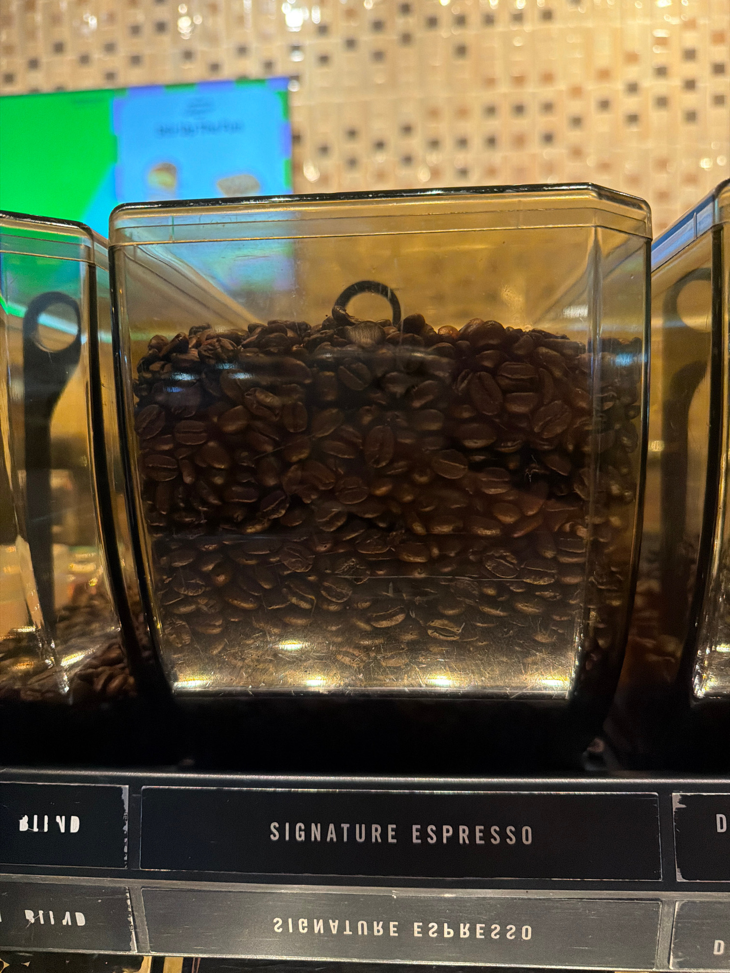 A clear plastic container filled with dark roasted coffee beans is positioned in front of a textured, tiled wall. To the right of the coffee beans, a black vertical display panel is visible. This panel has multiple sections, each with white text. One section clearly reads "SIGNATURE ESPRESSO". Another section, to its right and slightly faded, appears to have the text "2168BE220". Further down, the text "BLIND" is visible on one section, and below it, "RT|AD". The overall setting appears to be indoors, possibly a cafe or a retail environment in Wichit, Thailand, given the context. The lighting suggests it could be daytime, with artificial light highlighting the metallic sheen on some of the surfaces. No people are visible in the image.
