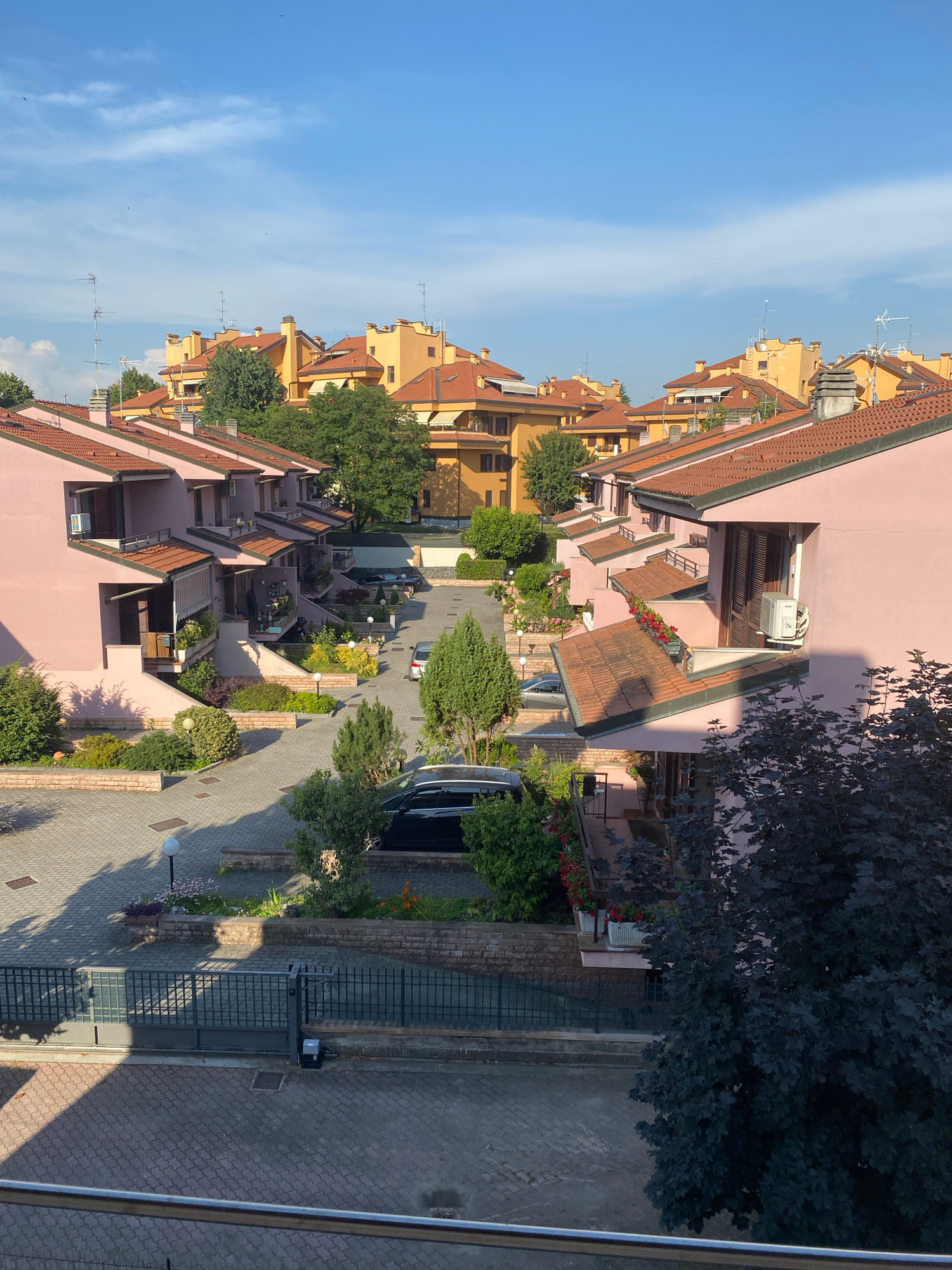 The image depicts a residential complex with multiple attached houses, characterized by pale pink facades and terracotta tile roofs. The houses are arranged in rows, with a central paved driveway and parking area. Greenery, including trees and manicured bushes, is interspersed throughout the complex. Some houses feature balconies with potted plants and flowers. Air conditioning units are visible mounted on the exterior walls of some residences. The sky is blue with scattered clouds, suggesting a clear day. A dark-colored car is parked on the driveway, and another vehicle is partially visible in the background. A black metal fence with an automatic gate is situated in the foreground. The architecture and landscaping suggest a suburban or urban residential setting in an Italian context.