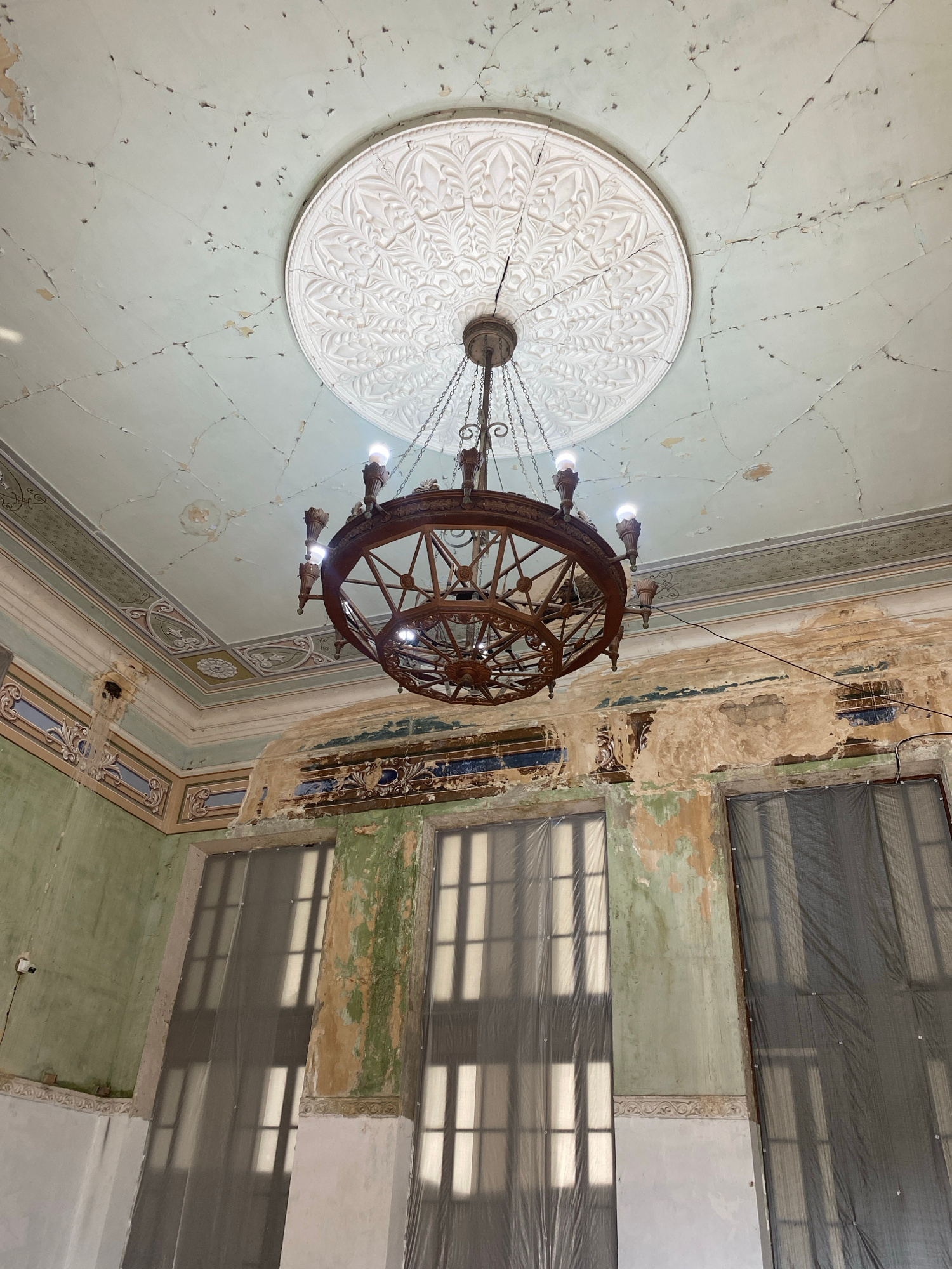 The image shows the interior of a room in Psakhara, Abkhazia. A large, ornate wooden chandelier hangs from a cracked, pale green ceiling adorned with a decorative plaster medallion. The walls are plastered and painted in shades of green, with sections of peeling paint and exposed plaster. Intricate decorative molding runs along the top of the walls, featuring floral and geometric patterns. Large windows, covered with translucent gray sheeting, cast patterned shadows on the walls and floor. The overall impression is one of decay and disrepair, with evidence of past grandeur. No people are visible in the image.