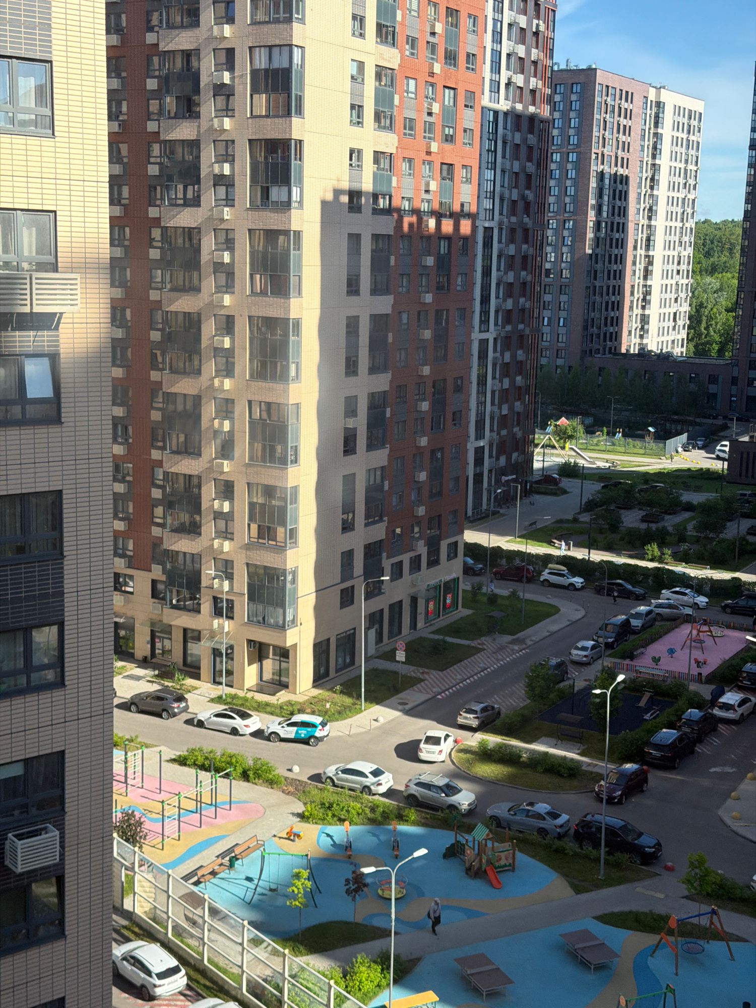 The image depicts a residential complex in Moscow, Russia, featuring multiple high-rise buildings with modern architecture. The buildings are primarily constructed with brick and glass facades, exhibiting variations in color from light beige to red and gray. The complex includes a children's playground area with swings, slides, and colorful surfacing, as well as parking areas with numerous cars. The scene is bathed in sunlight, casting shadows from the buildings, and a clear blue sky is visible. There are no obvious signs of activity or interaction between people, with only a few individuals sparsely scattered throughout the scene, one of whom is walking near a playground. The overall impression is one of a contemporary urban living environment.