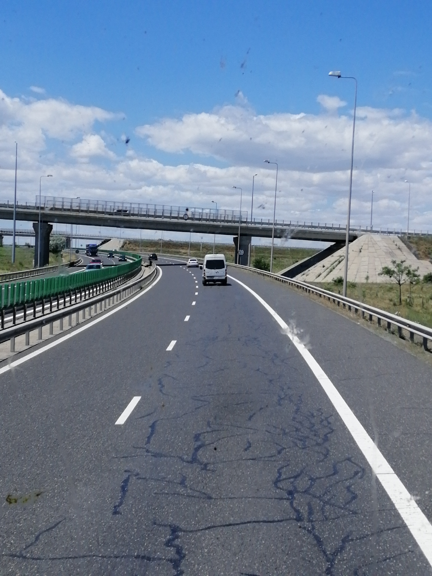 The image shows a highway under a bright blue sky with scattered white clouds. The highway has multiple lanes separated by white lines and a green guardrail on the left. An overpass with concrete supports and railings is visible in the background, with cars and a truck driving on it. Several vehicles, including a white van, a black car, and a dark-colored truck, are on the highway in the foreground. The road surface shows some cracking and dark patches. The environment appears to be a rural or semi-rural area with dry grass and a few scattered trees visible beyond the highway. The presence of a highway overpass and the general landscape suggest a location on the outskirts of a city. Given the provided context, this scene is likely located on the A4 motorway (Autostrada Soarelui) near Constanța, Romania, which connects Constanța to Bucharest and features numerous overpasses and elevated sections.