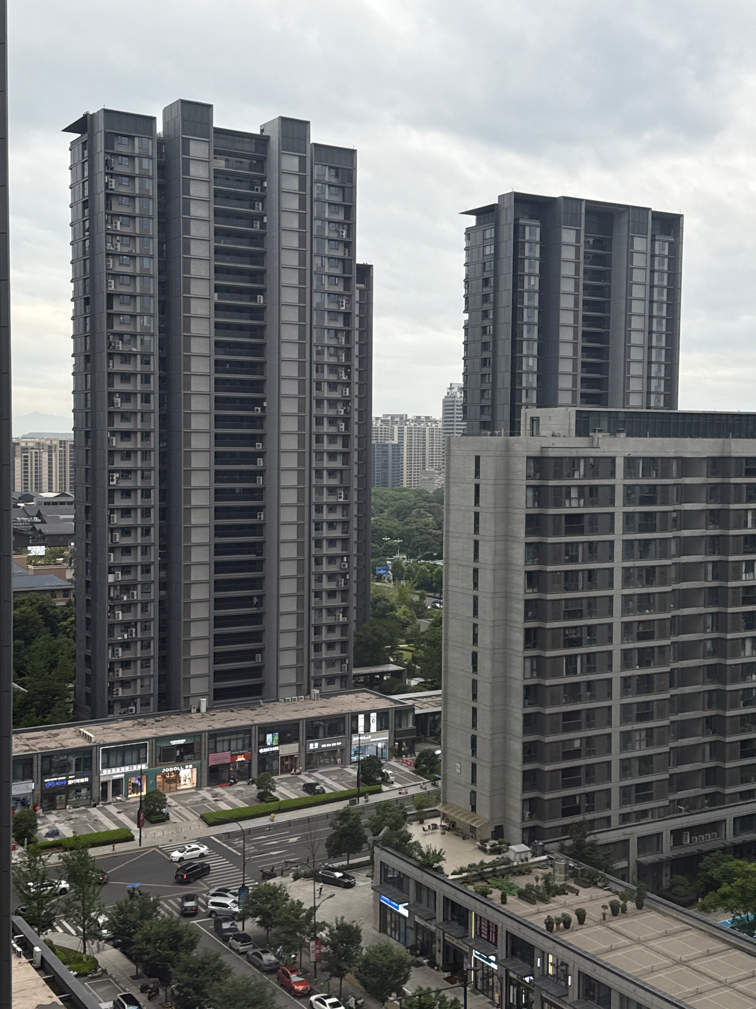 This is a vertical, eye-level, medium shot capturing a cityscape in Yiwu, China, on a cloudy day. The scene is dominated by modern high-rise apartment buildings, characterized by their dark grey facades and repetitive window patterns. In the foreground, a low-rise building with commercial storefronts is visible, featuring illuminated signs and a street with parked cars and moving traffic. A pedestrian crossing is present, with a few individuals walking. The background reveals more buildings and lush green trees, suggesting a well-developed urban environment. The sky is overcast, with diffused light, casting a uniform, soft illumination over the scene.