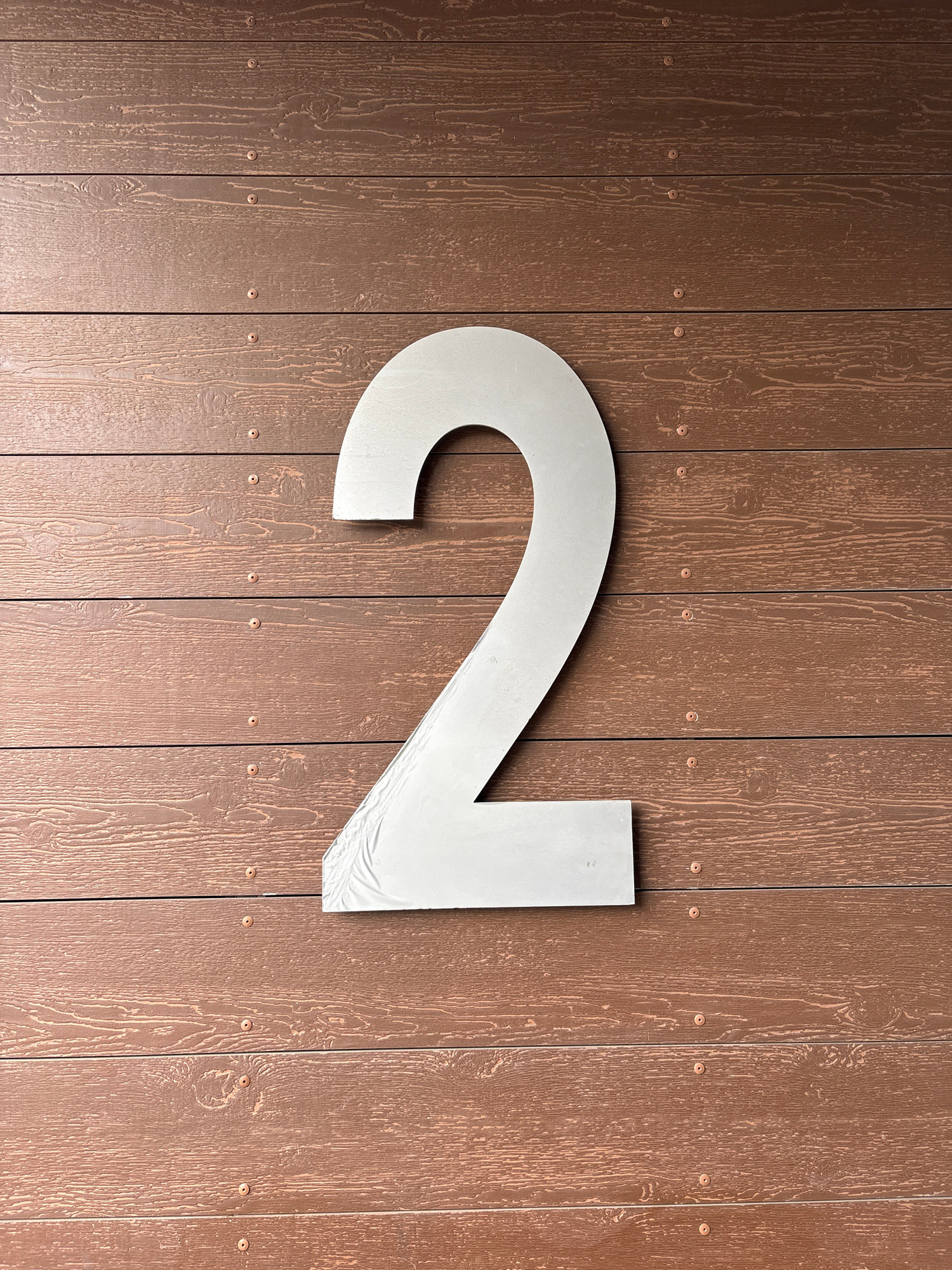 This image features a large, silver, three-dimensional number "2" mounted on a vertical wooden plank surface. The planks are stained a medium brown with a visible wood grain texture, and they are fastened with small, circular, copper-toned rivets or nails. The number "2" is positioned centrally, dominating the frame, and appears to be made of metal, possibly aluminum or steel, with a matte finish. The texture on the number suggests it might be a cast or fabricated piece. The lighting indicates it is daytime, casting soft shadows that highlight the dimensional aspects of the number. The background is consistent throughout, suggesting a uniform surface, possibly an exterior wall of a building or a fence. There is no text visible in the image, other than the number "2" itself. The scene is static and focuses solely on the number and its mounting surface. The location context suggests this could be a house number or a marker in Mytishchi, Russia, but there are no specific architectural details or geographical cues to confirm this definitively beyond the provided context. The overall impression is one of simple, modern signage against a rustic backdrop.