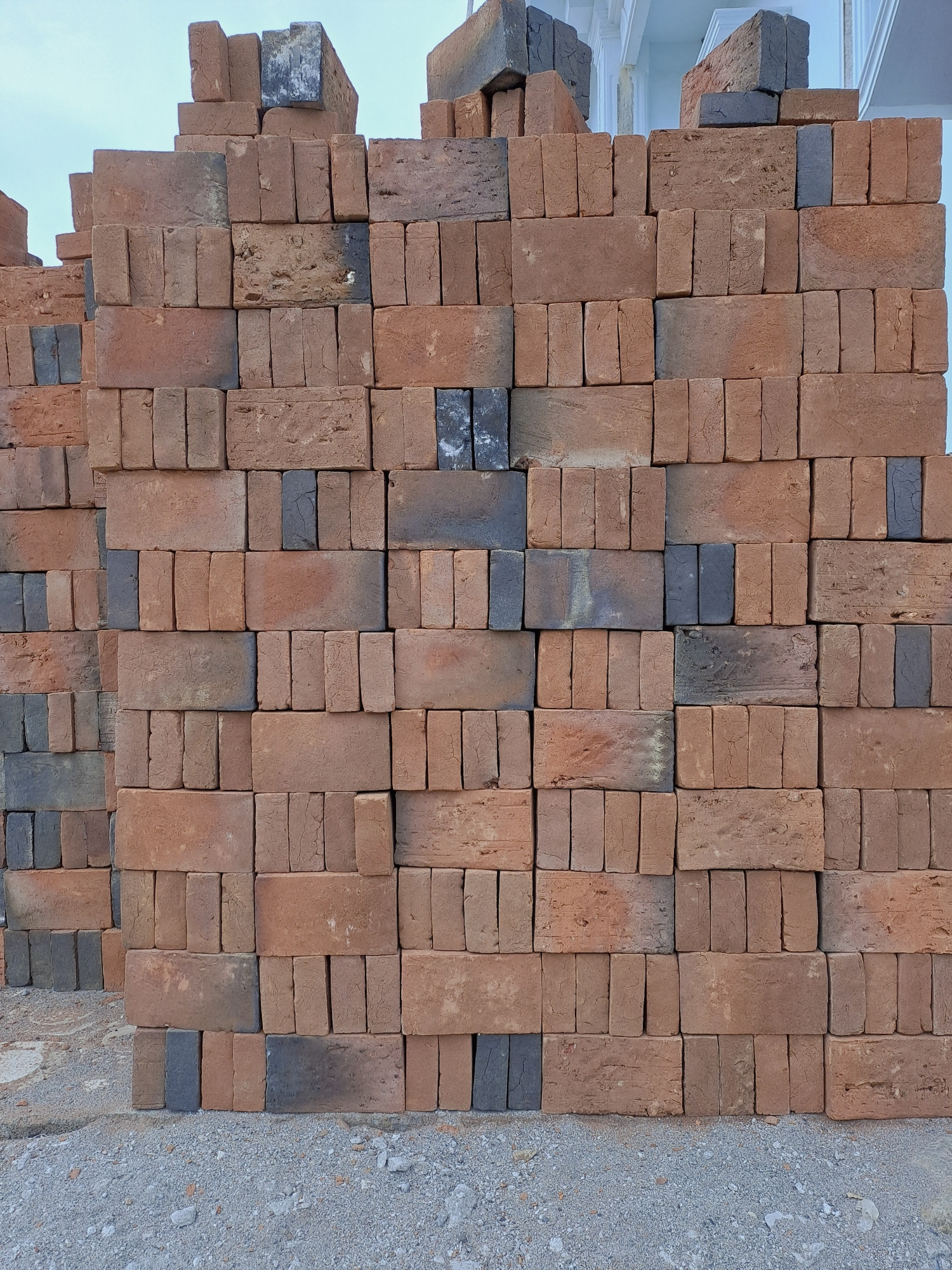 A large stack of red and grey bricks is piled high against a light-colored building facade in Lhokawe Teungoh, Indonesia. The bricks are of varying sizes and conditions, with some appearing worn and chipped. They are stacked in an uneven, somewhat haphazard manner, with several rows reaching well above eye level. The ground beneath the stack is covered in gravel and small stones. The sky is visible at the top of the frame, appearing pale blue and overcast. The scene depicts a construction or storage area where building materials are being kept.