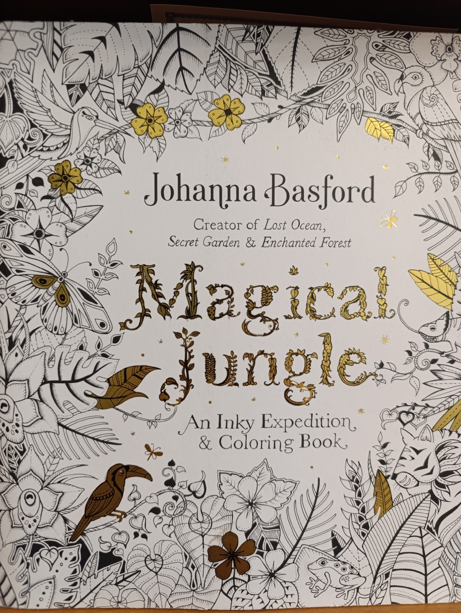 The image is a close-up of the cover of a coloring book titled "Magical Jungle" by Johanna Basford. The cover features intricate black and white line drawings of various jungle flora and fauna, with accents of metallic gold ink. There are illustrations of birds, butterflies, a toucan, a frog, and a tiger. The text "Johanna Basford", "Creator of Lost Ocean, Secret Garden & Enchanted Forest", and "Magical Jungle, An Inky Expedition & Coloring Book" are prominently displayed in elegant fonts. The background is white, and the overall aesthetic is detailed and whimsical. The lighting suggests it's an indoor setting, likely a bookstore or a home. The image is an eye-level shot of the book cover.