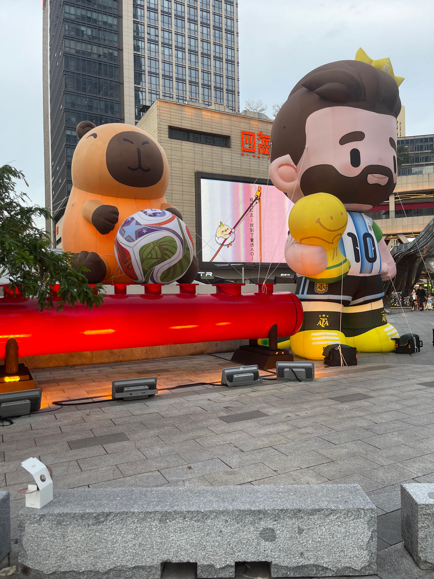In Panyu District, China, a large-scale outdoor display features two inflatable cartoon characters. On the left, a tan bear-like character sits holding a colorful soccer ball. To its right stands a larger inflatable figure dressed in a soccer uniform with the number 10, holding a golden trophy with a smiley face. The figures are placed in front of a building with a large LED screen displaying a cartoon image. A red, illuminated structure runs horizontally in front of the inflatables, with lighting equipment on the ground. The scene is set on a paved plaza with granite benches in the foreground.