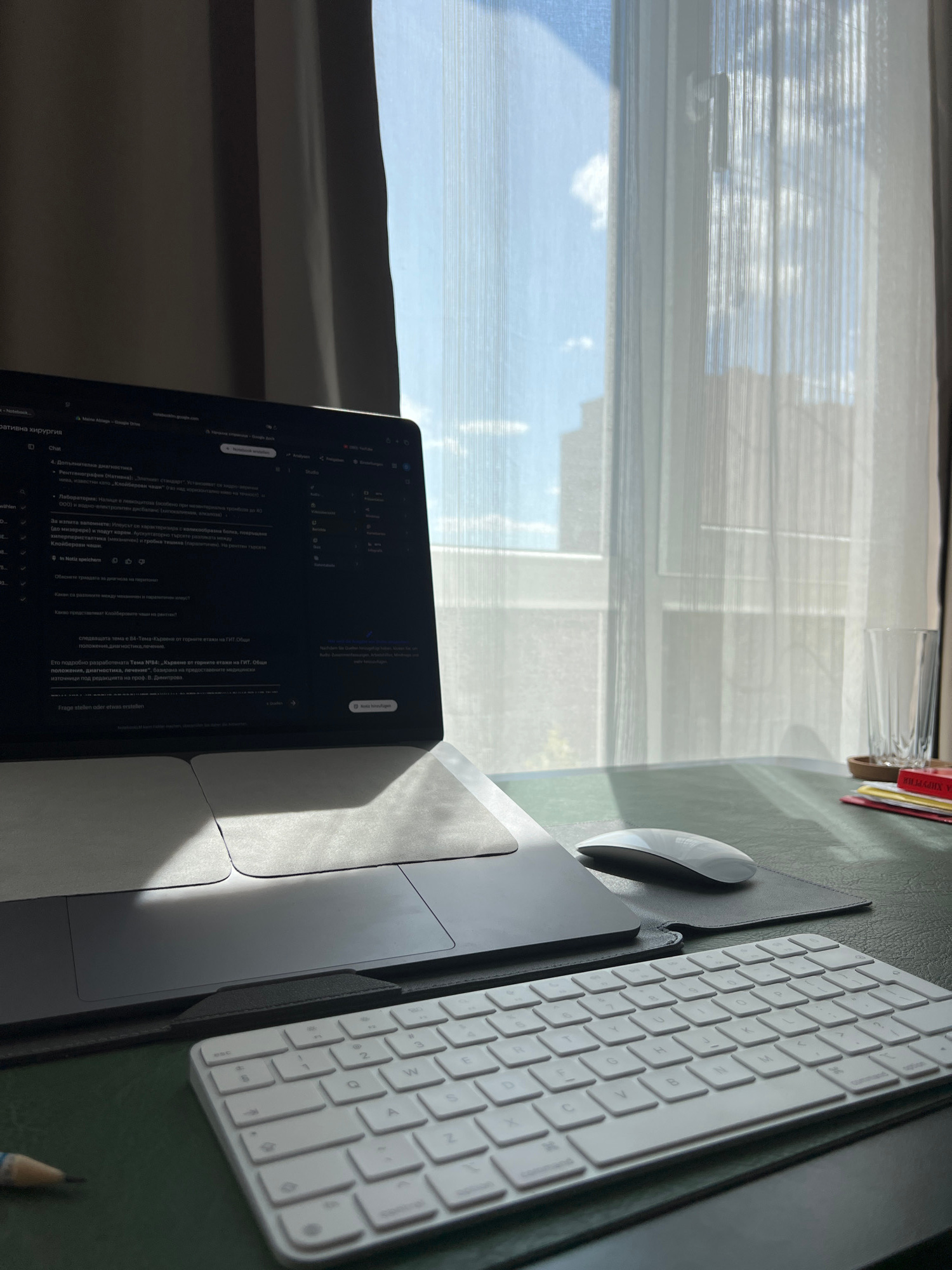 This image captures a desk setup bathed in natural light, suggesting it's daytime. The scene appears to be indoors, possibly a home office or study, given the presence of a laptop, a keyboard, and a mouse on a dark green desk pad. The laptop is open and displaying text, likely a document or website, with some Cyrillic script visible, indicating a potential setting in a region where this language is used, such as Bulgaria. The text on the screen includes medical terminology like "рентгенография" (radiography) and "хирургия" (surgery), suggesting the content relates to medicine.
To the left of the desk, sheer white blinds cover a window, allowing diffused sunlight to illuminate the scene and cast soft shadows. Through the blinds, a hint of blue sky and clouds is visible, suggesting clear weather outside. A close-up view of the blinds in the cropped images reveals horizontal stripes and the faint outlines of buildings or structures beyond.
On the desk, a white keyboard and a white mouse are positioned next to the laptop. A dark-grey laptop sleeve or case is also visible, partially covering the trackpad area of the laptop. In the background, a few items are partially visible, including what looks like a glass and some colored papers or folders, adding to the impression of a workspace. The overall mood is calm and focused, typical of a setting where work or study is taking place. The visible text on the laptop screen provides a specific clue about the subject matter being reviewed, pointing towards medical education or research.