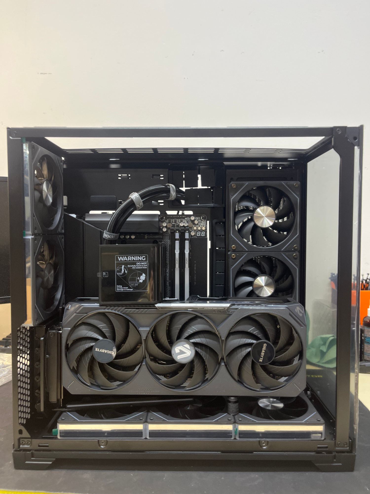 A high-end gaming computer is assembled inside a clear-sided PC case. The computer features a large graphics card with three fans, a liquid cooling system with tubes, and multiple case fans. The components are primarily black and silver, with some RGB lighting visible. The computer is placed on a dark, flat surface, likely a desk or table, in an indoor setting with a plain white wall in the background. There are no people or animals present in the image, and no geographical markers that would indicate a specific location in Kyiv, Ukraine.