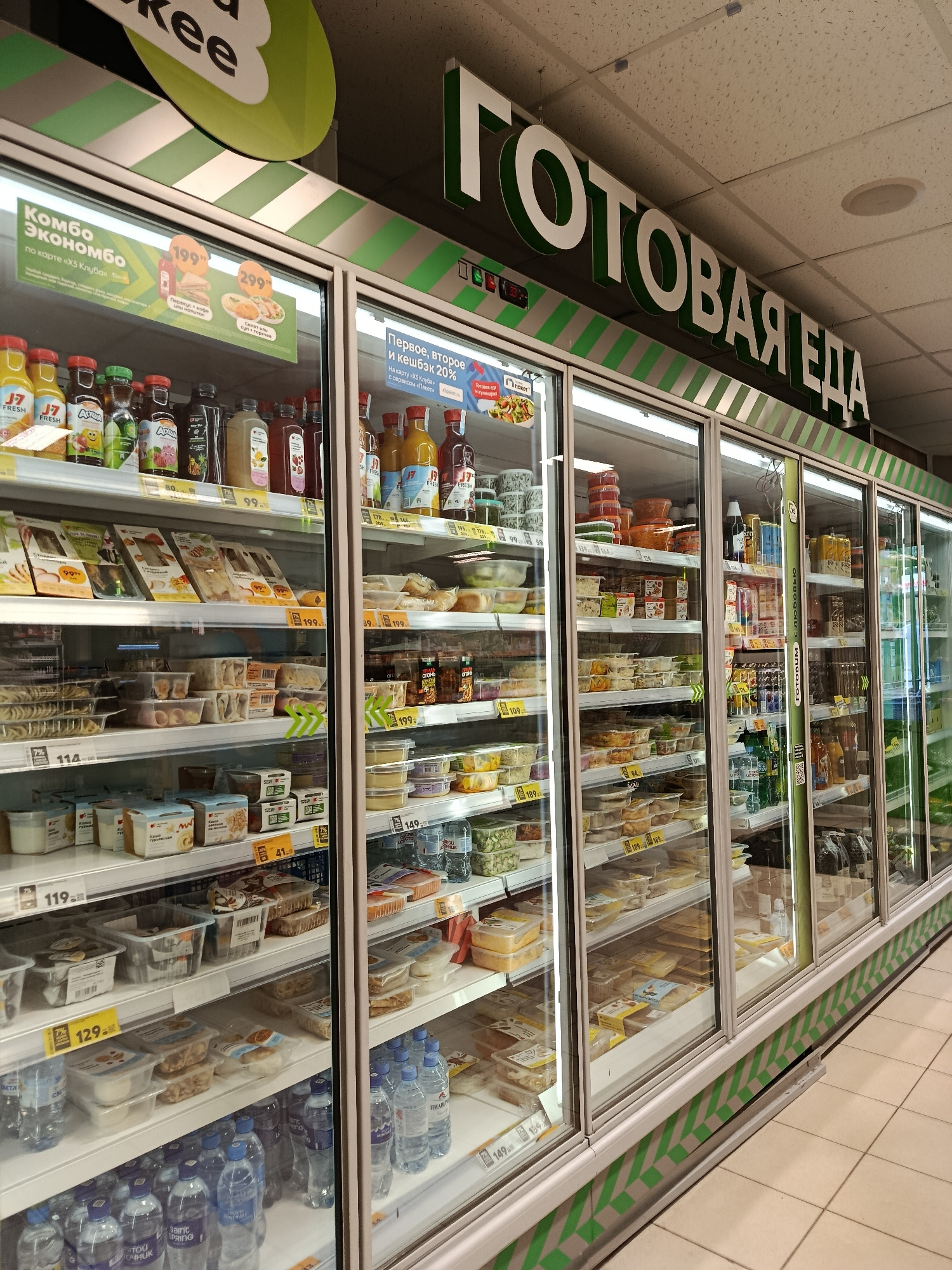 This media file shows a refrigerated display case in a supermarket. The display case is filled with a variety of pre-packaged food items, including meals, salads, and beverages. The products are arranged on multiple shelves, and many have visible price tags.
The scene is brightly lit, suggesting an indoor setting, likely during daytime operating hours of the store. The overall impression is one of a well-stocked grocery store offering ready-to-eat food options. The prominent text "ГОТОВАЯ ЕДА" (Ready-to-eat food) in large green and white letters above the display case clearly indicates the nature of the products offered. Other visible text includes promotional messages such as "Комбо Экономбо" (Combo Economy), "Первое, второе и кешбэк 20%" (First course, second course, and 20% cashback), and brand names like "J7" and "АГУША".
The location is specified as Верея, Russia, providing geographical context for the supermarket. There are no people visible in the image, and the focus is entirely on the products and the store's display. The atmosphere appears clean and organized, typical of a modern retail environment.